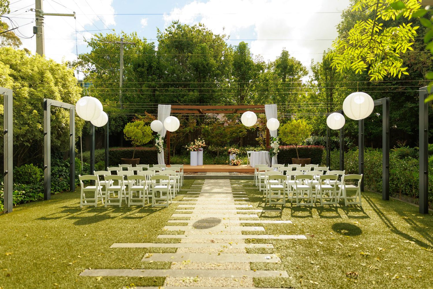 Sunlit outdoor wedding venue with white folding chairs, paper lanterns, and a floral altar.
