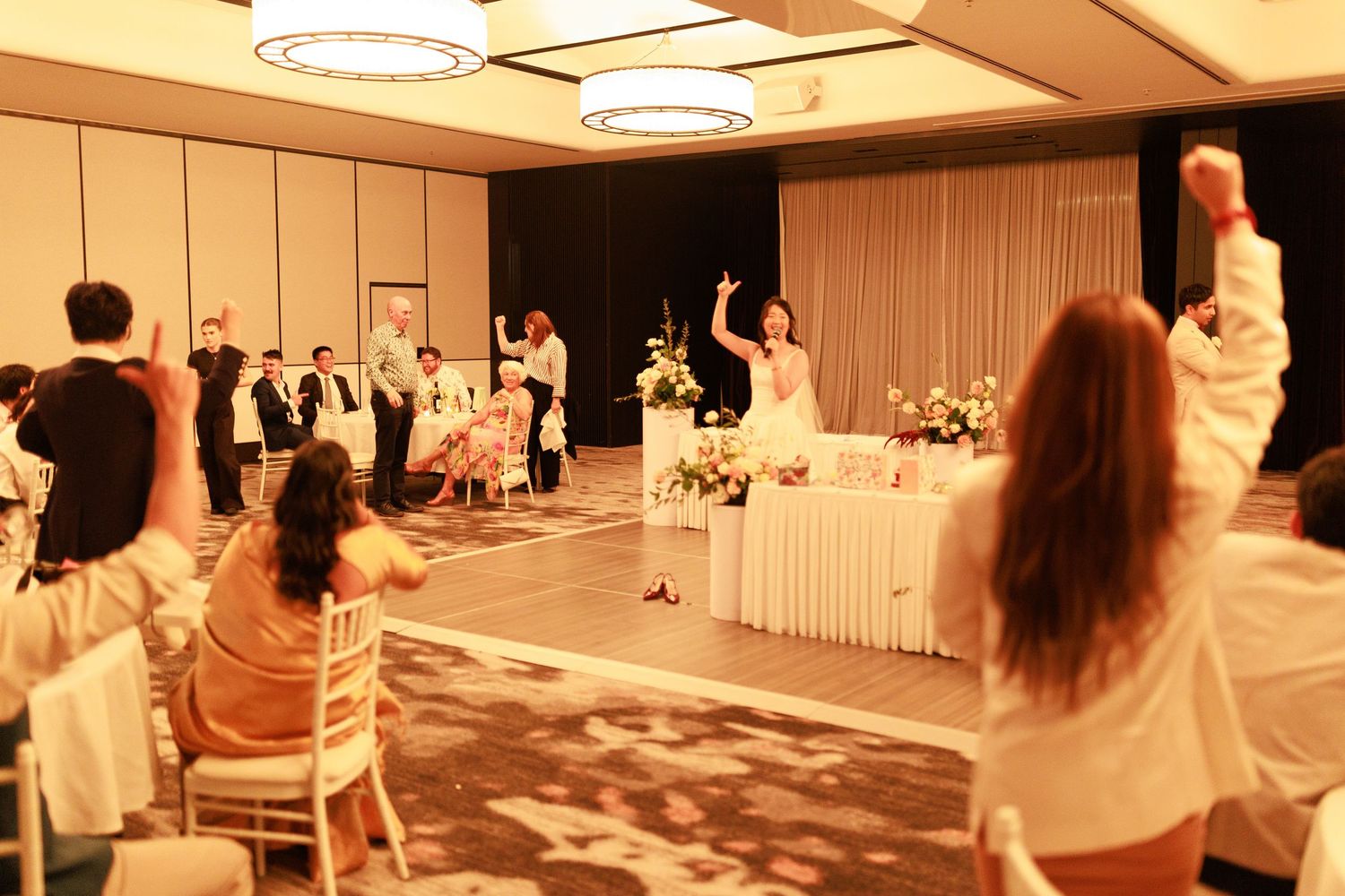 Wedding reception toast with bride and guests raising hands in elegant hotel ballroom celebration.