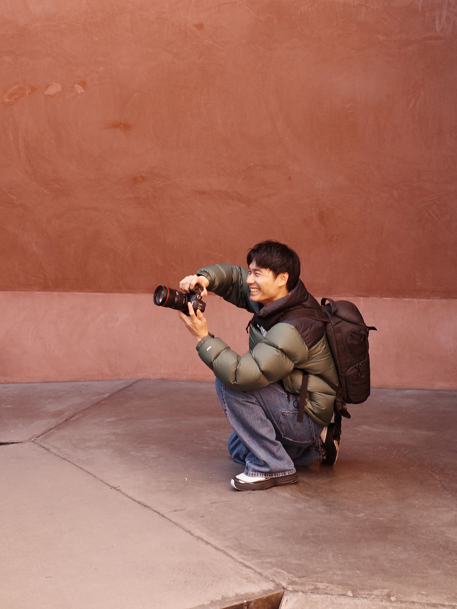 Asian male photographer smiling while crouching with camera against red textured wall.