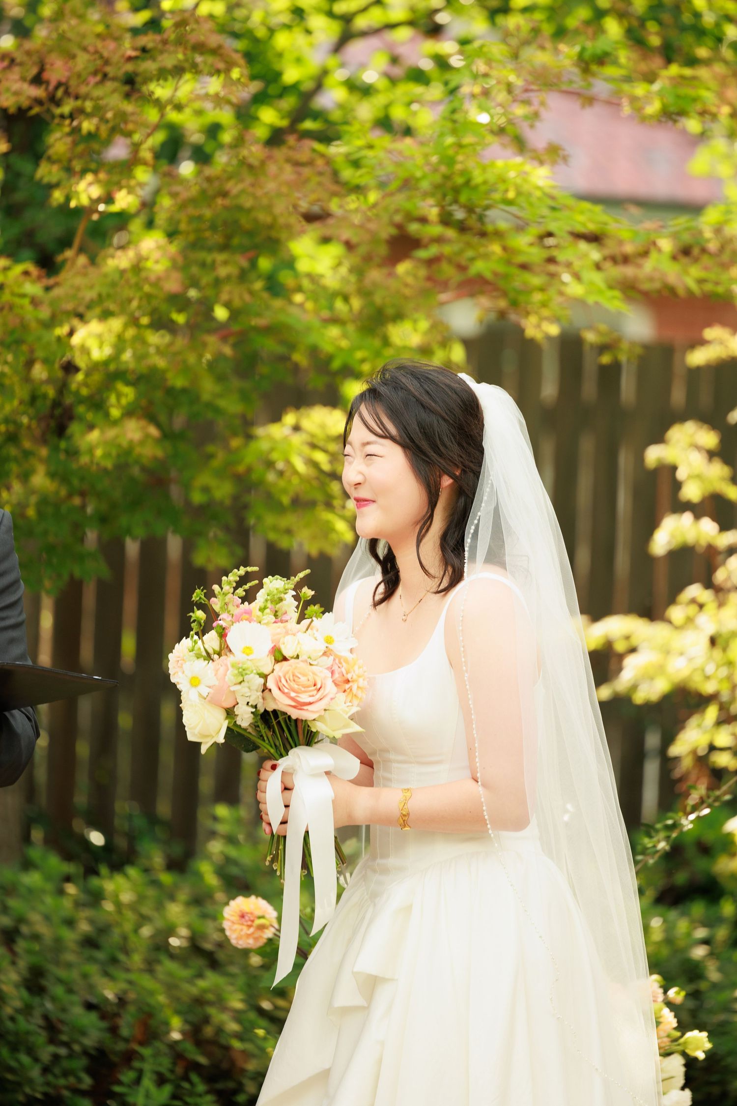 Smiling Asian bride in white gown and veil holds peach floral bouquet outdoors in garden.