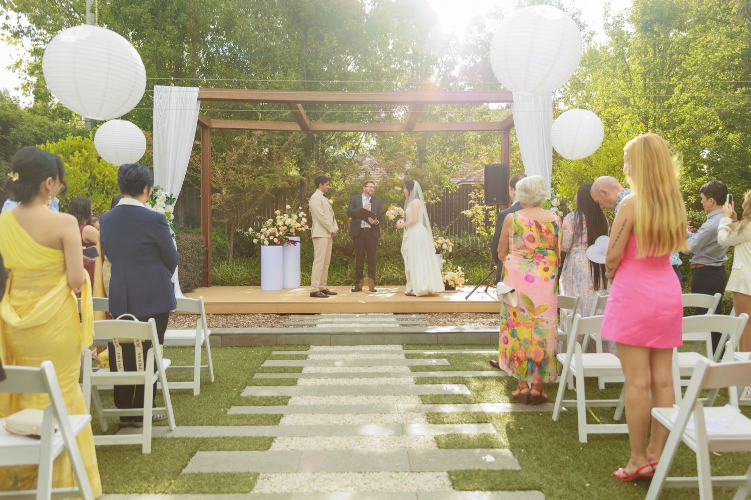 Outdoor wedding ceremony in a sunlit garden with guests seated in white chairs facing the altar.