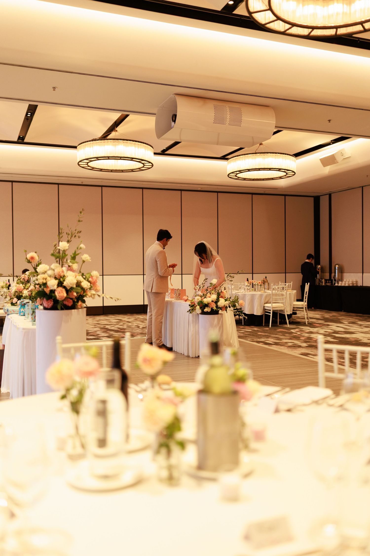 Wedding venue setup with staff arranging floral centerpieces on tables in elegant ballroom.