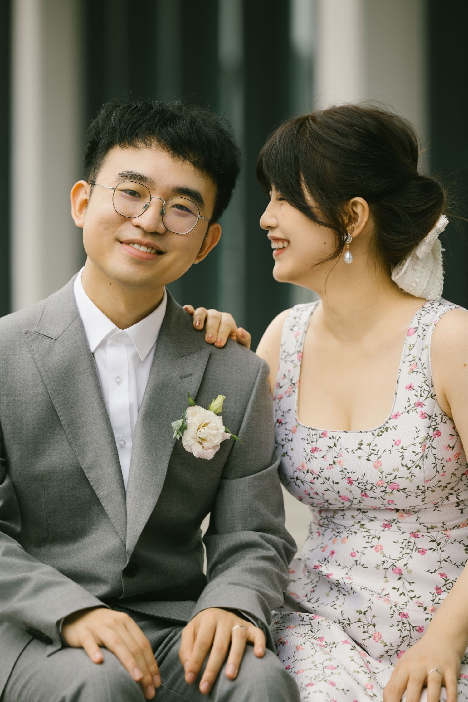 Smiling Asian couple in wedding attire sit together outdoors, groom in gray suit with boutonniere.