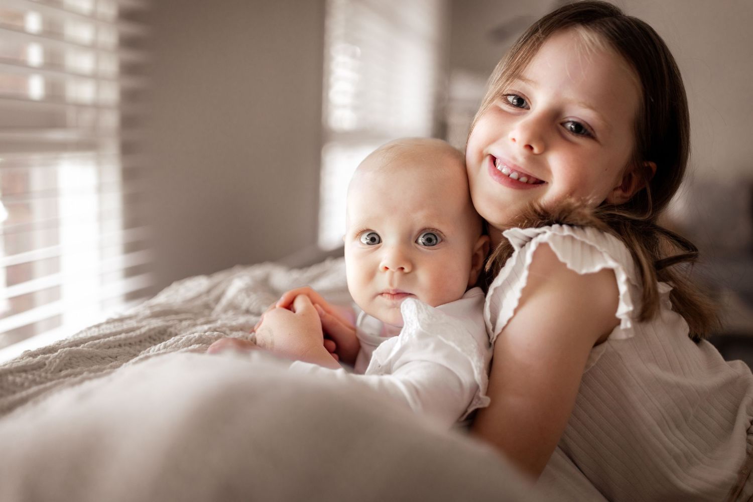 Older sister lovingly hugging baby sibling on a soft white bed in natural light.