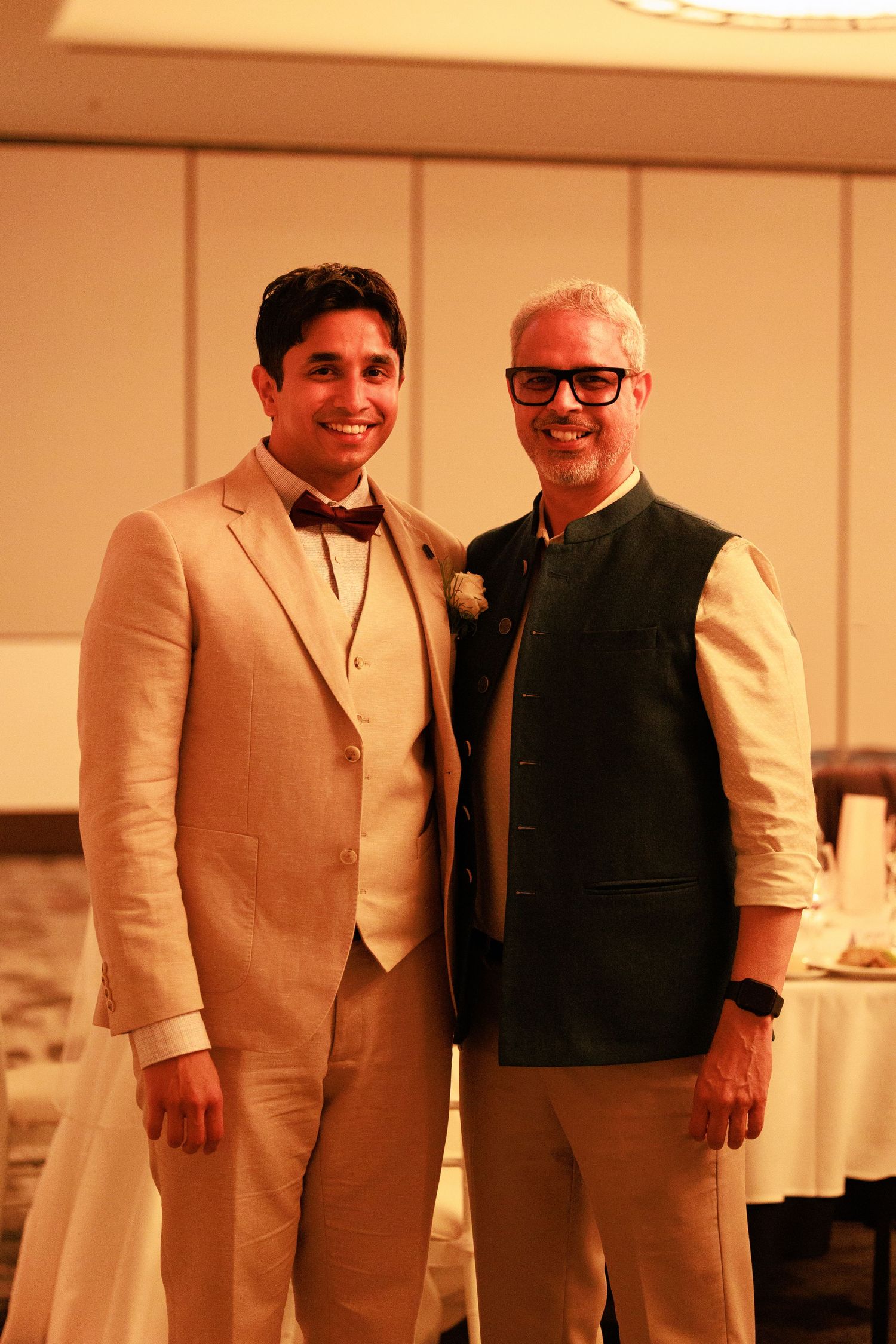 Two men smiling together at a wedding reception, one wearing a beige suit with bow tie.