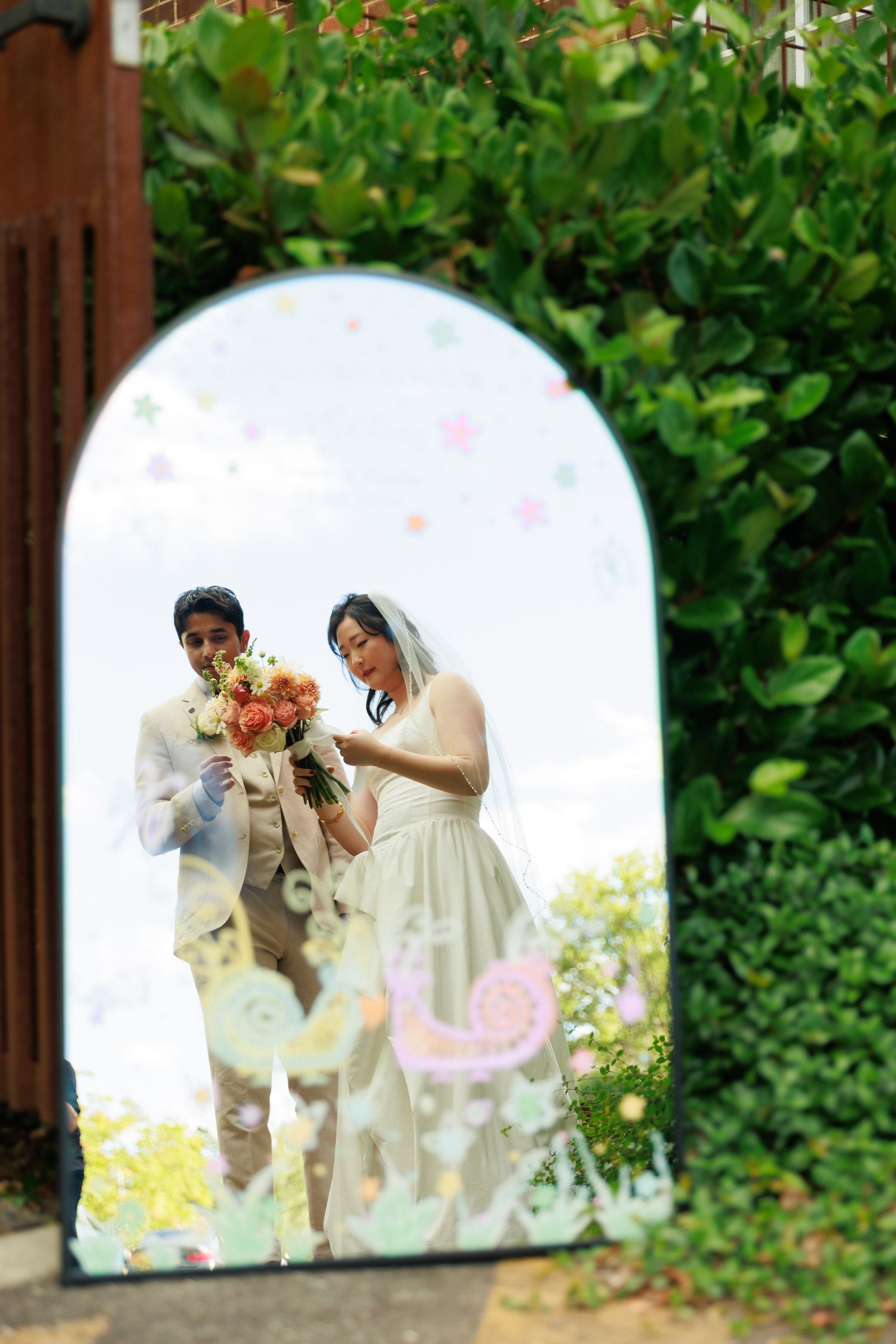 Wedding couple reflected in an arched decorative mirror surrounded by green hedges outdoors.