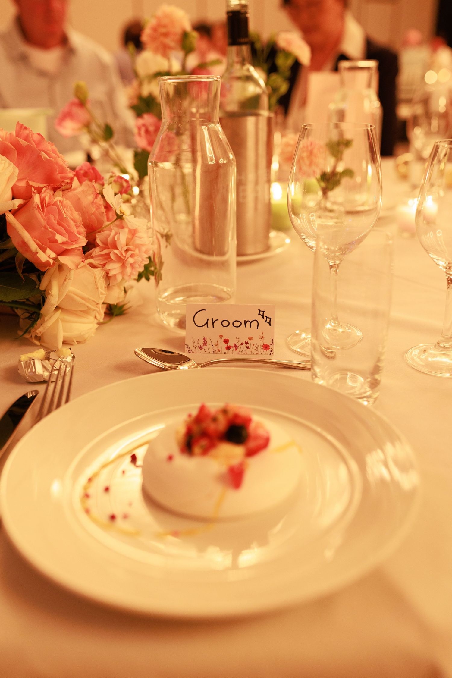 Elegant wedding reception table setting with groom place card, white plate, pink flowers and glassware.