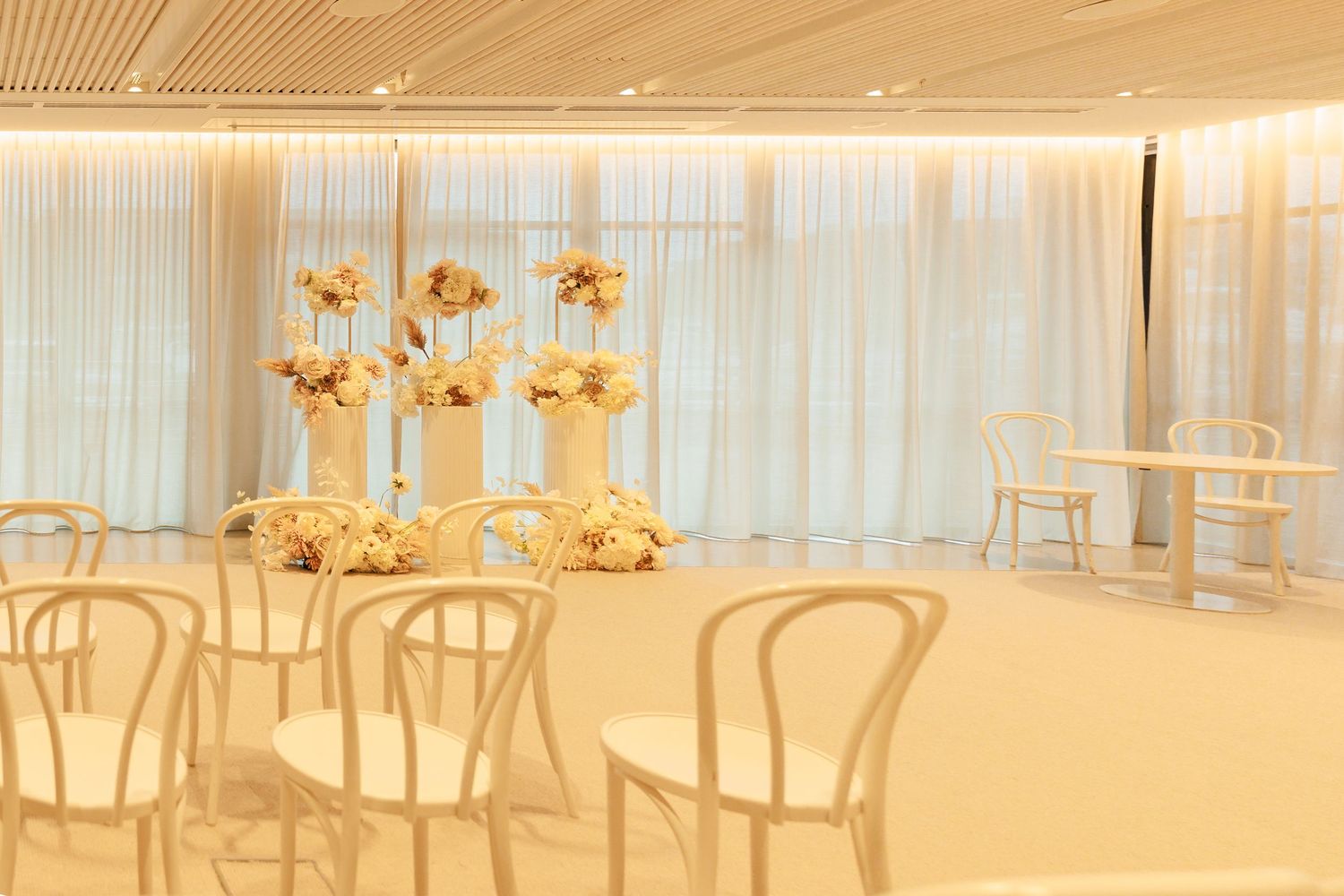 A bright minimalist wedding venue with white chairs, sheer curtains, and elegant tall dried floral centerpieces.