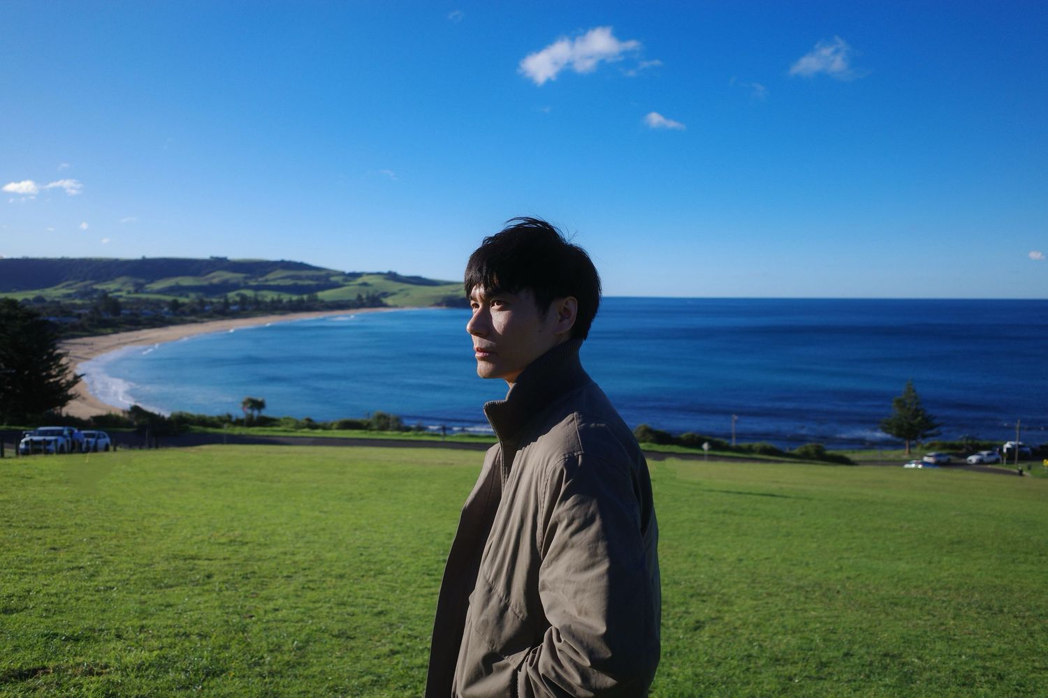 Young man standing on green hillside overlooking a stunning blue ocean bay on a sunny day.