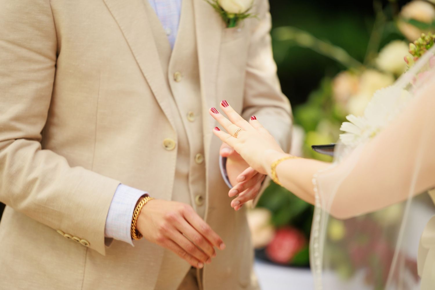 Bride with red nails places wedding ring on grooms finger during outdoor ceremony.
