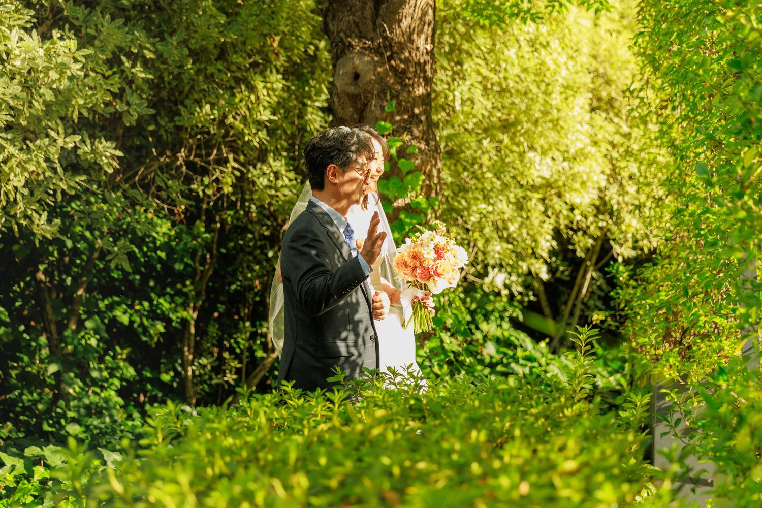 Bride and groom embracing among vibrant green foliage with a colorful bouquet in sunlit garden.