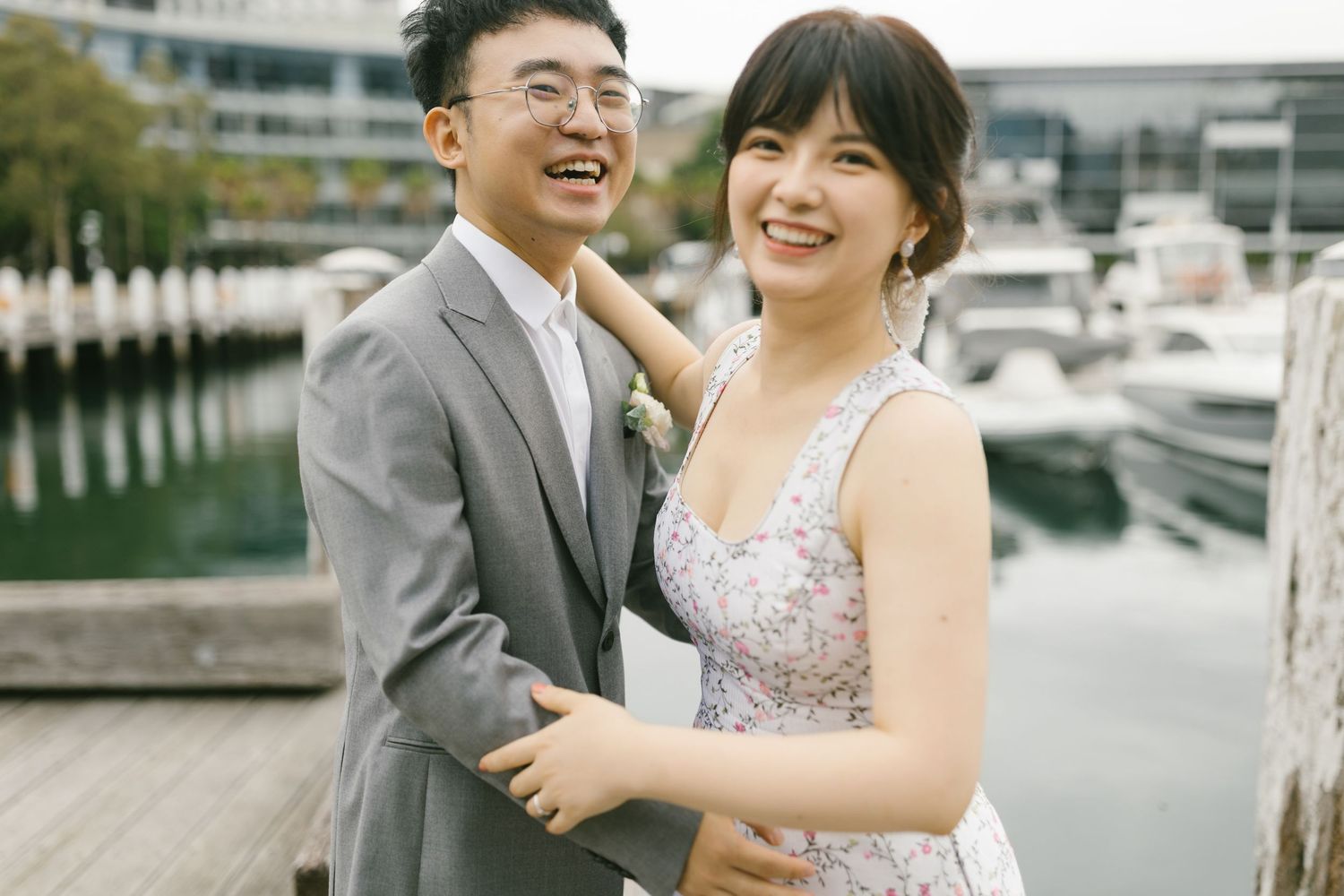 A happy Asian couple in wedding attire laughing together on a dock with boats in the background.