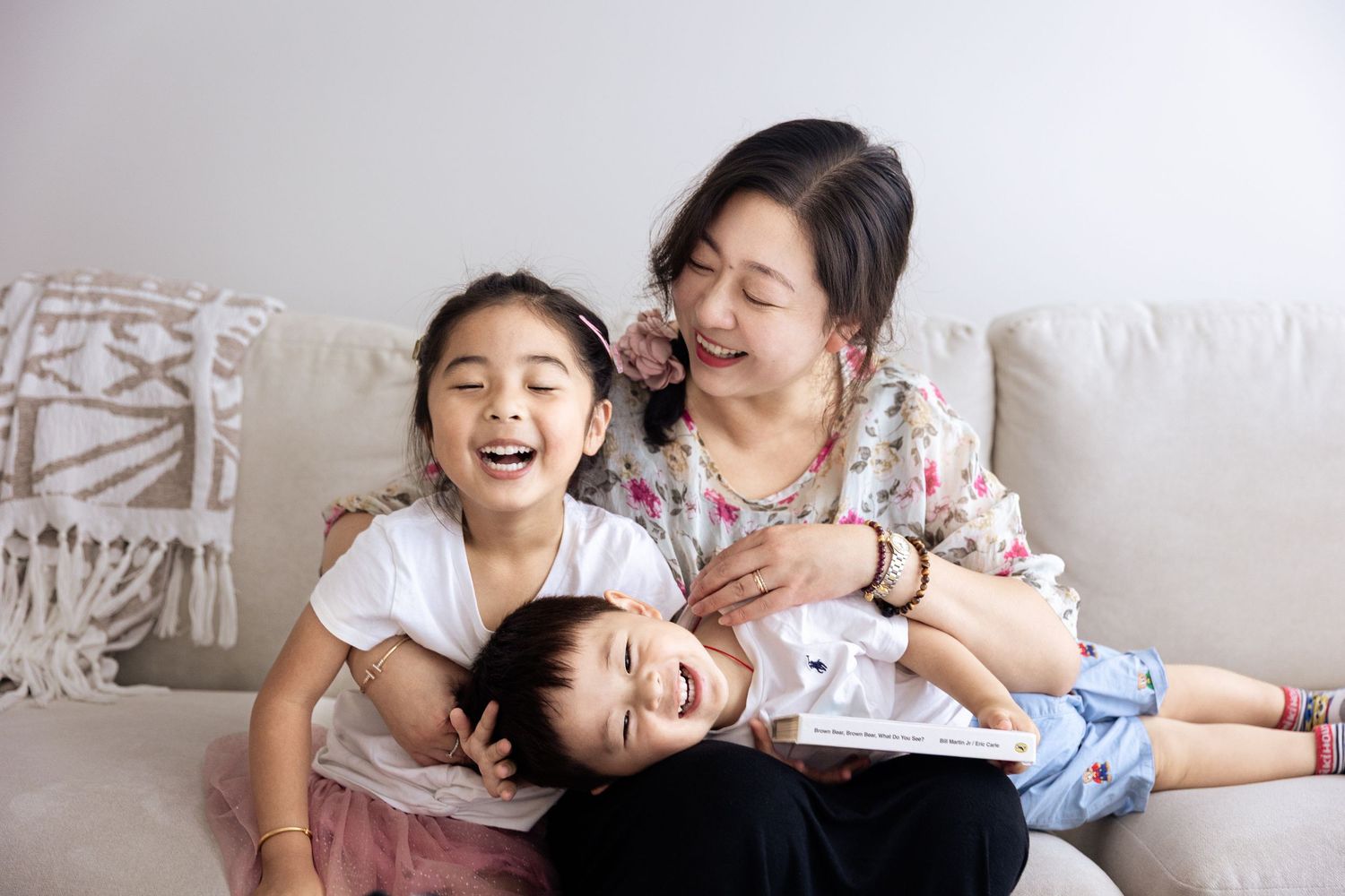 A joyful Asian mother and two children laughing together on a couch, sharing a fun family moment at home.
