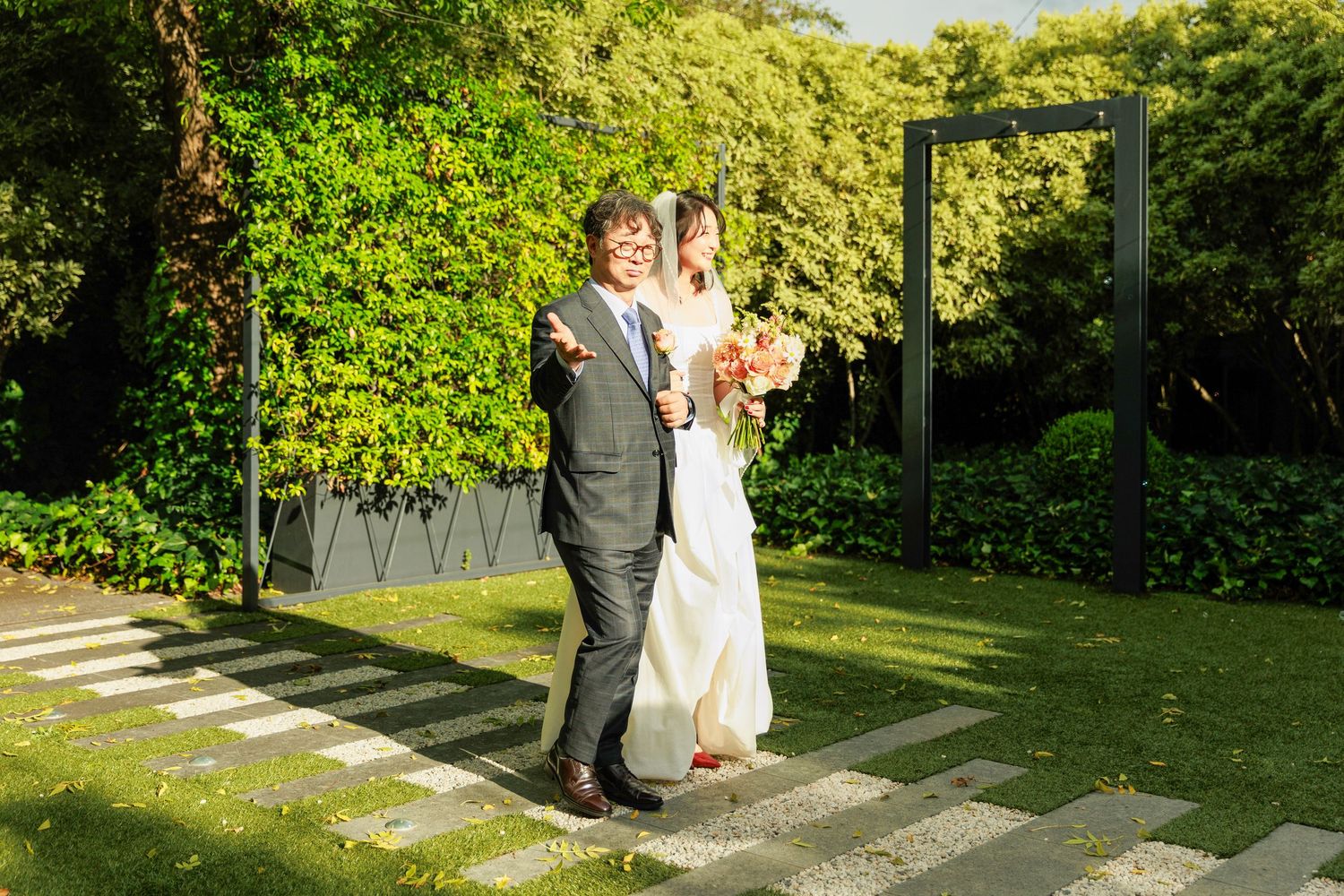 Bride and groom walking together on a garden path with lush green hedges and modern frame backdrop.