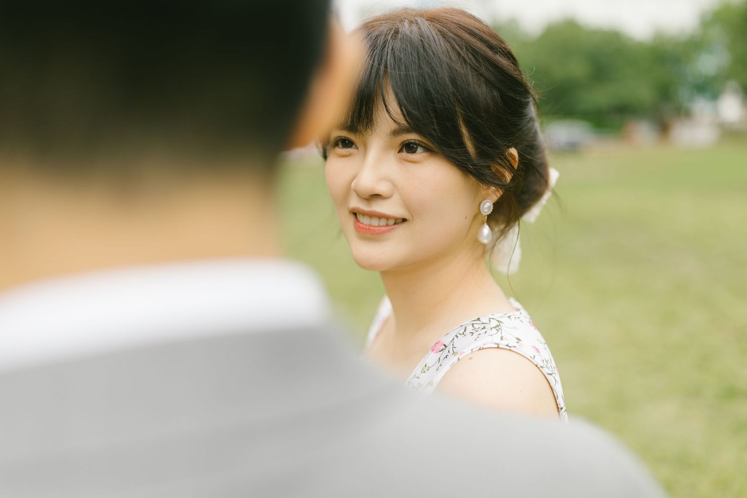 A smiling Asian bride in a white dress gazes softly over her shoulder at her partner outdoors.