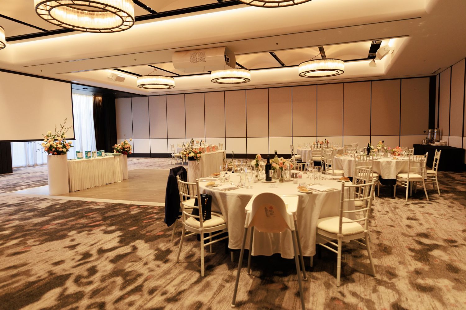 Spacious wedding reception ballroom with round tables, white linens, chiavari chairs and floral arrangements.