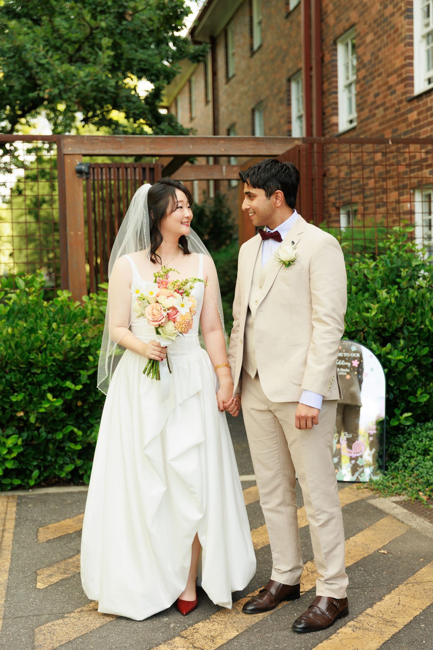 Bride and groom holding hands and smiling at each other outside a brick building with lush greenery.