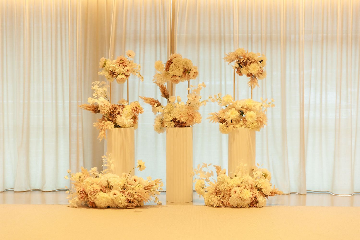 Three elegant dried floral arrangements in cream and beige tones displayed on white pedestals against sheer curtains.