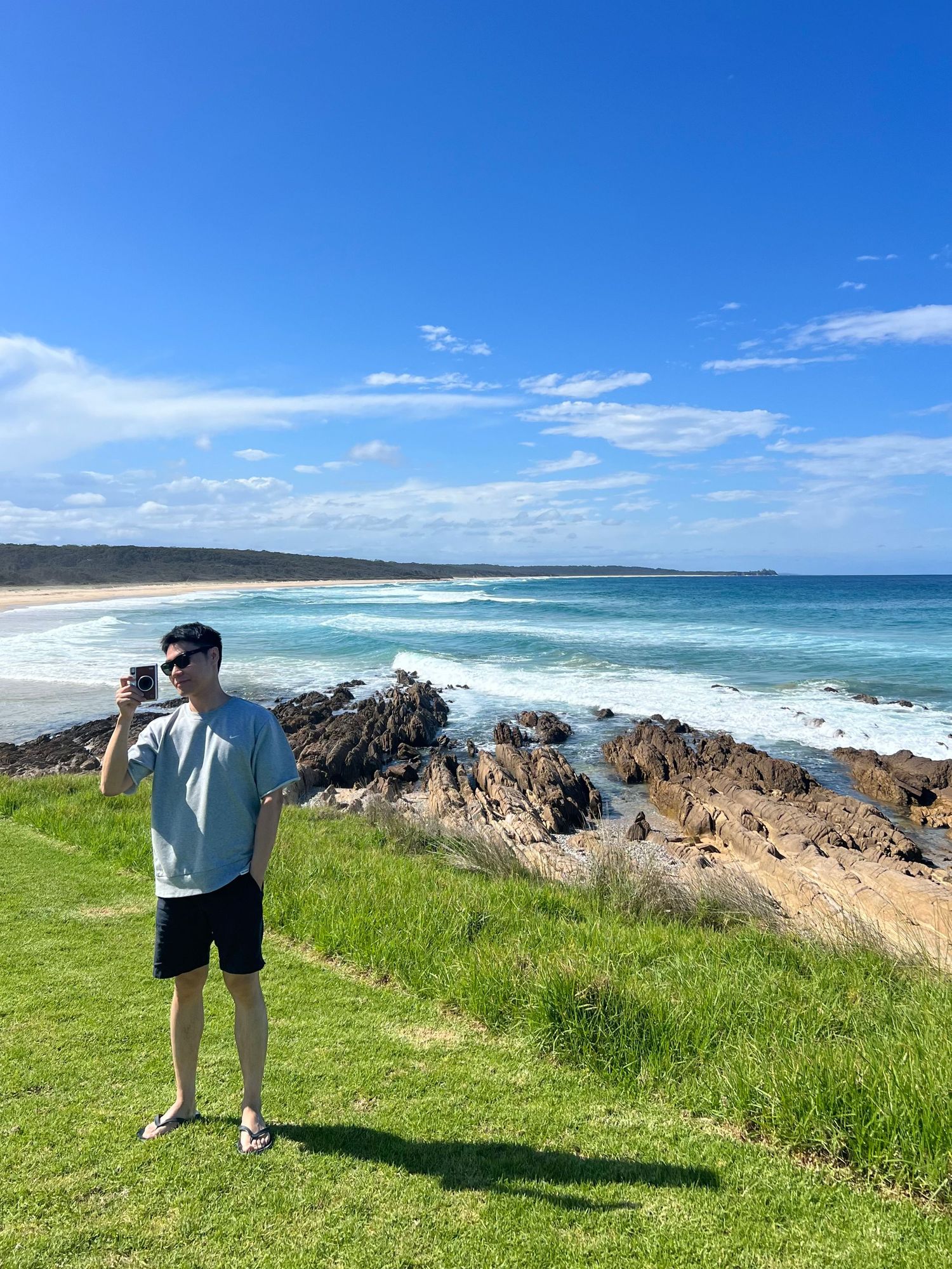 Man in casual outfit photographs stunning Australian coastal landscape with turquoise waves and rocky shoreline.