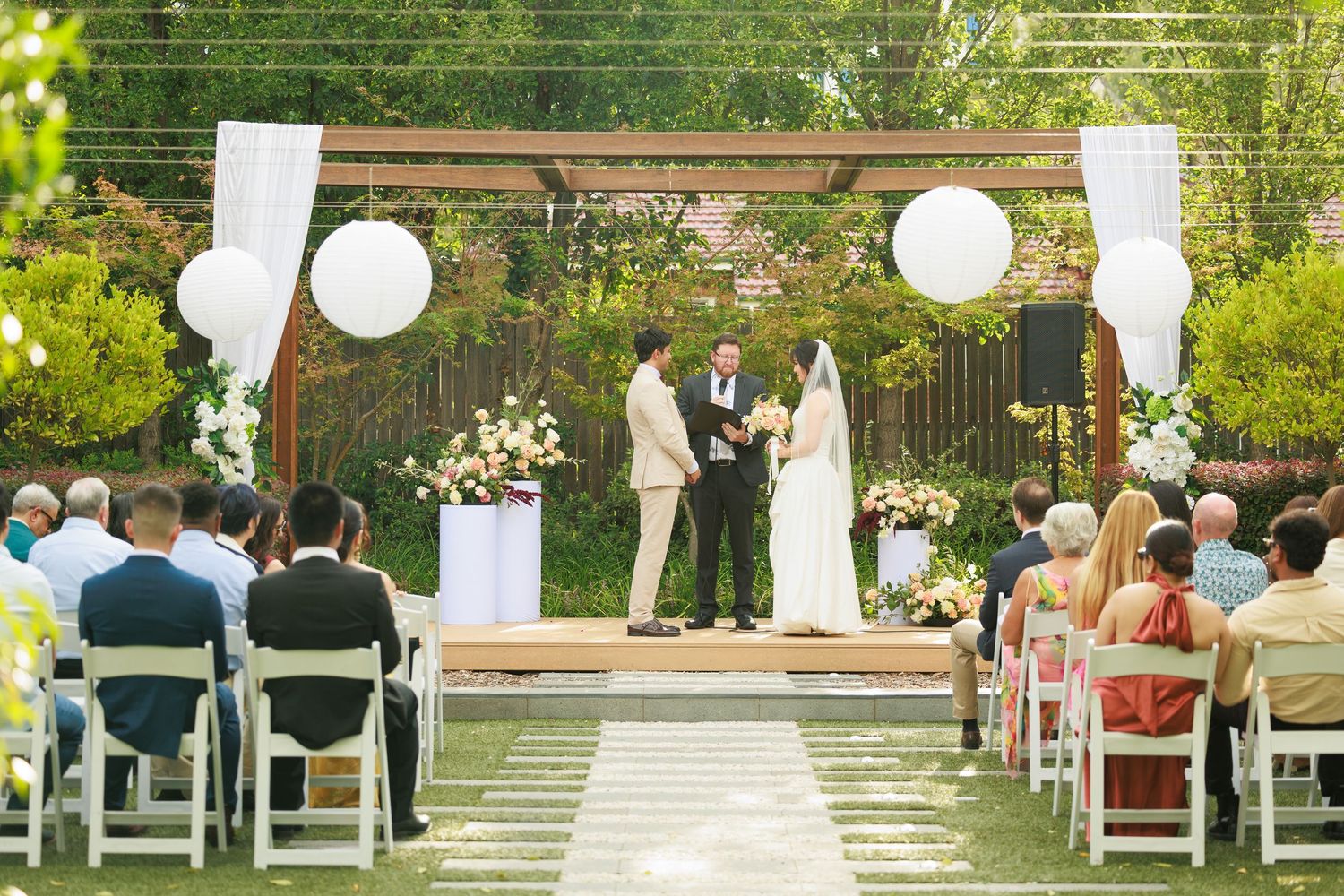 Outdoor garden wedding ceremony with couple at floral altar, white balloon decor, and seated guests.