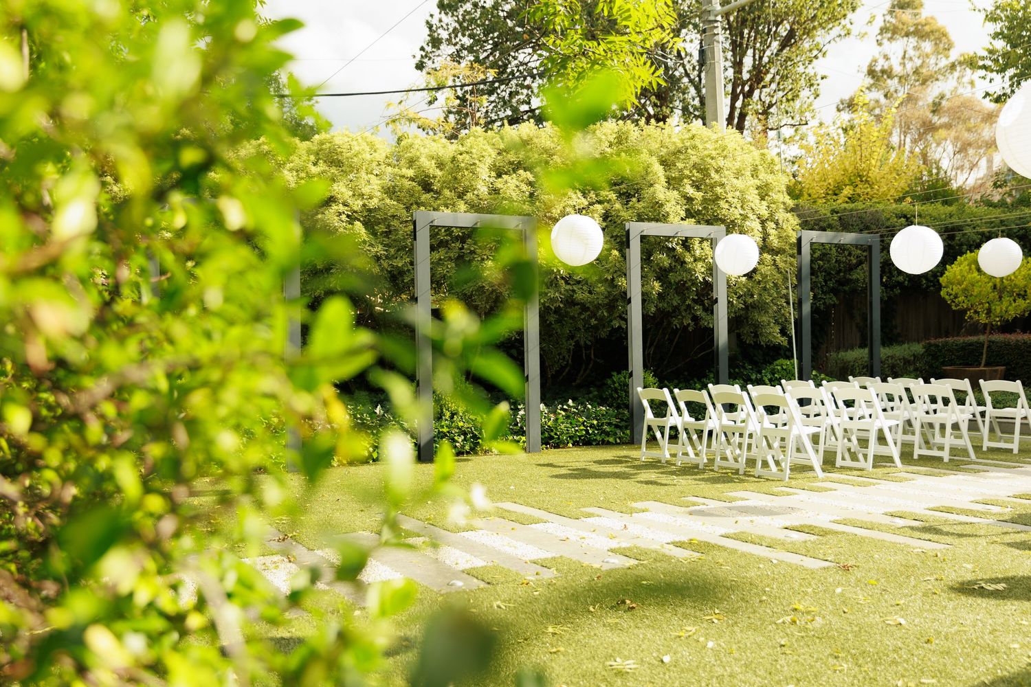 Elegant outdoor wedding ceremony setup with white chairs, paper lanterns, and lush garden backdrop.