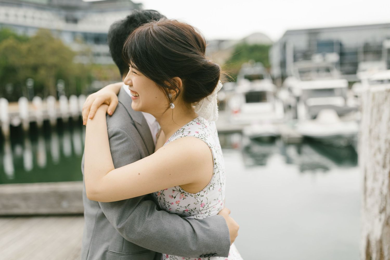 A bride in a floral dress shares a warm embrace with her groom on a waterfront dock.