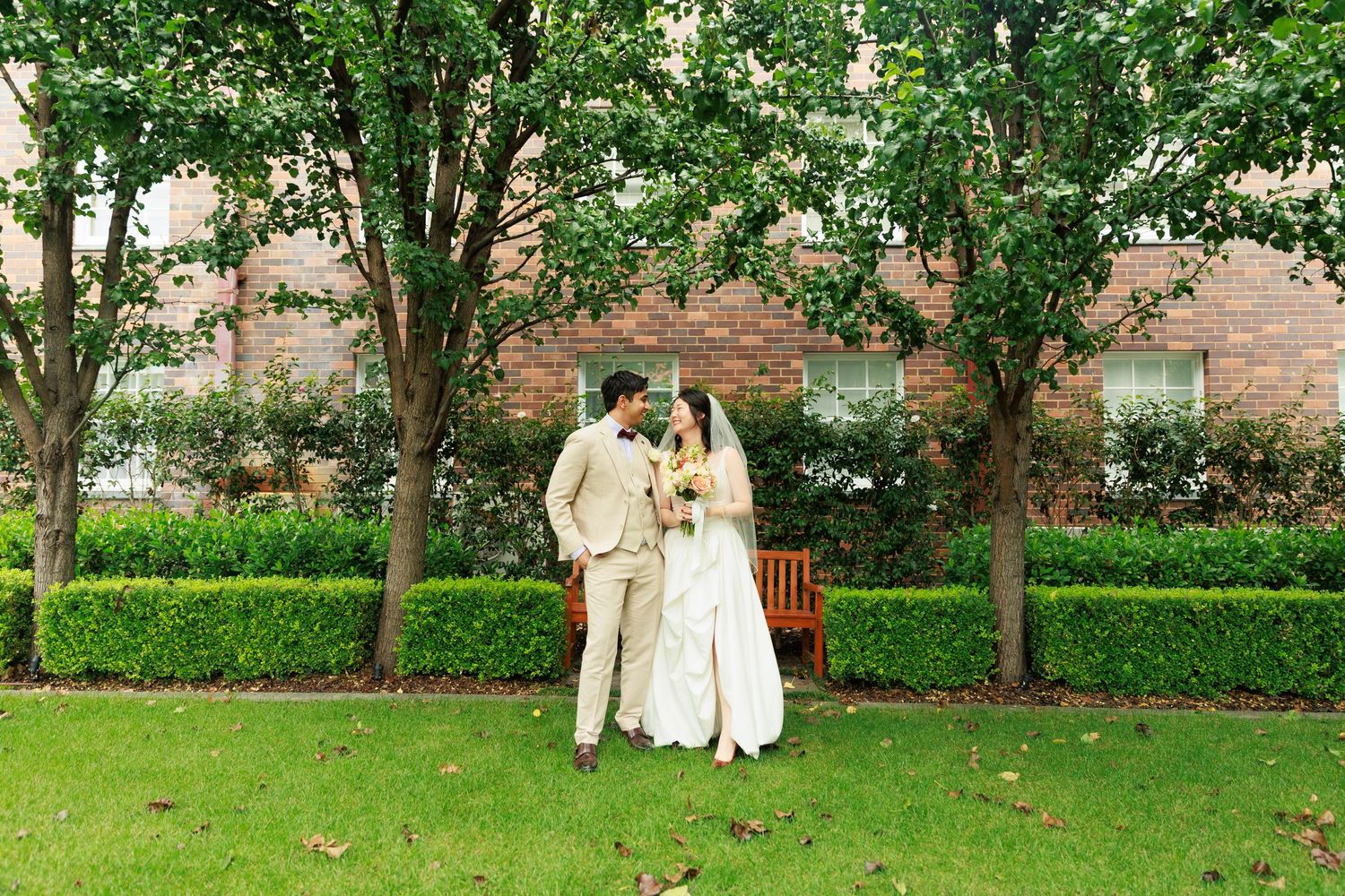 Bride and groom sharing a romantic moment on a green lawn with trees and brick building backdrop.