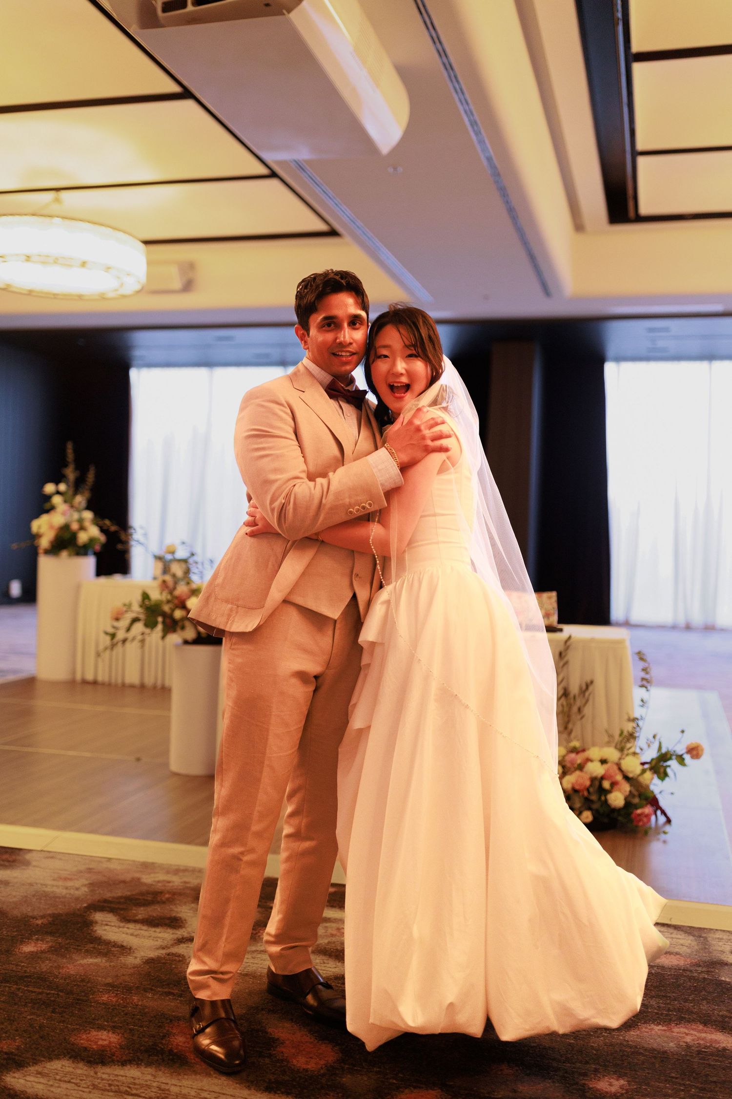 Newlywed couple embraces and laughs joyfully in decorated wedding reception ballroom.