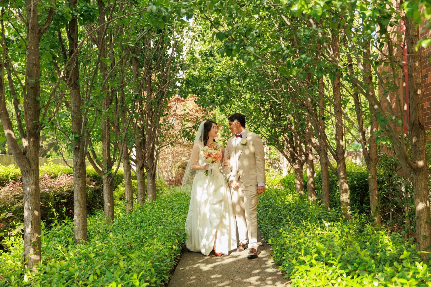 Bride and groom walking hand in hand through a lush green tree-lined garden path on their wedding day.