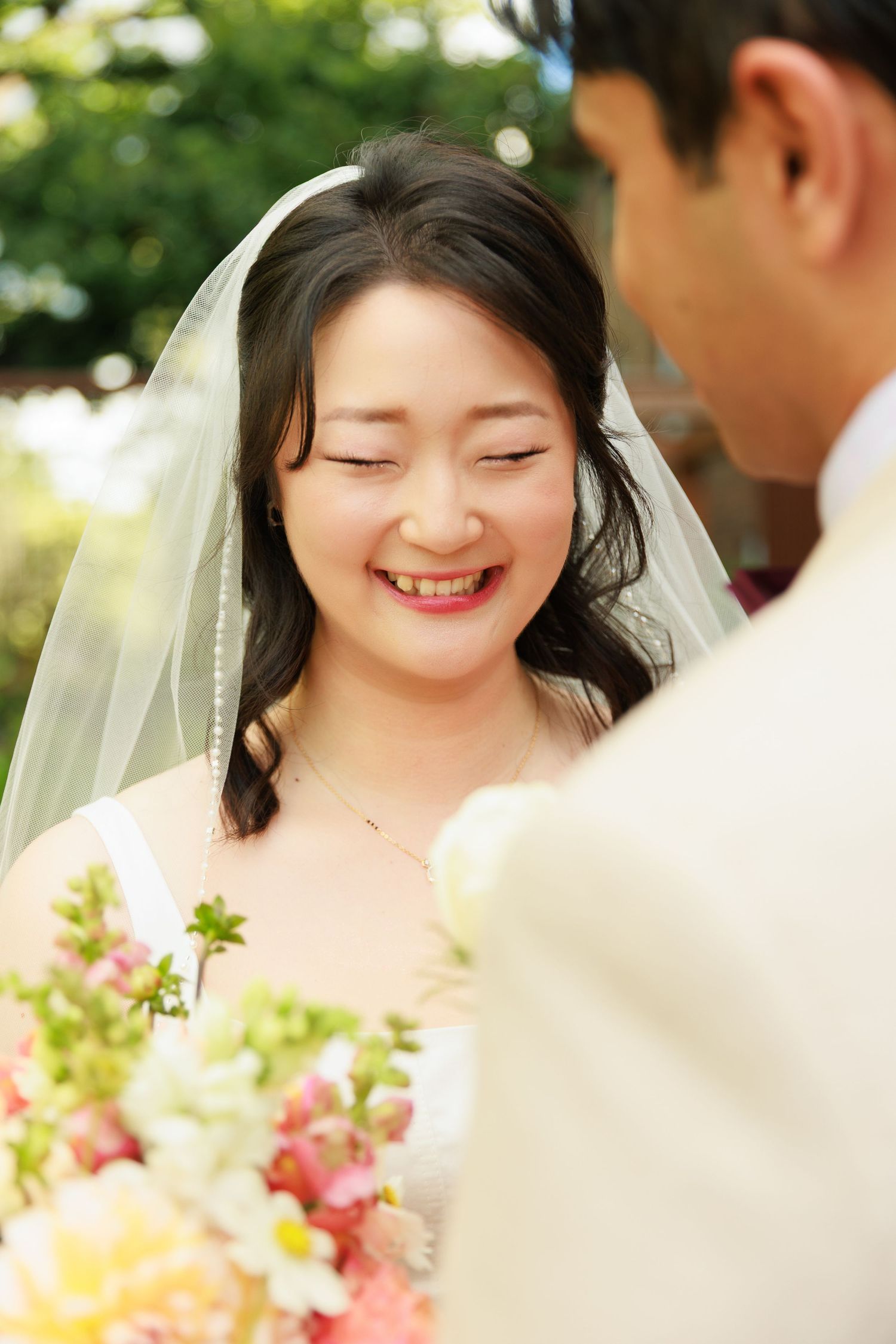 Joyful bride in white veil smiling warmly while holding a colorful bouquet outdoors in sunlight.