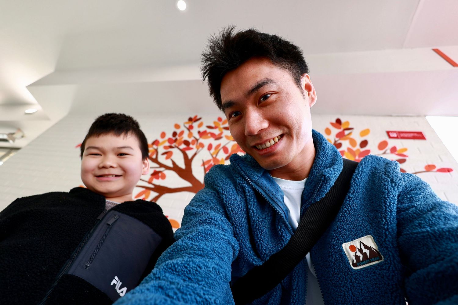 Two young Asian men taking a selfie indoors near a colorful autumn tree mural.