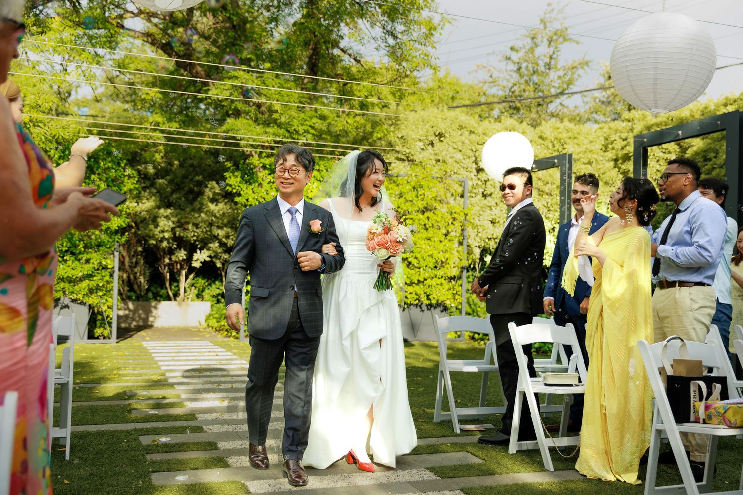 Bride walks down outdoor wedding aisle with escort, guests seated on white chairs in lush garden setting.
