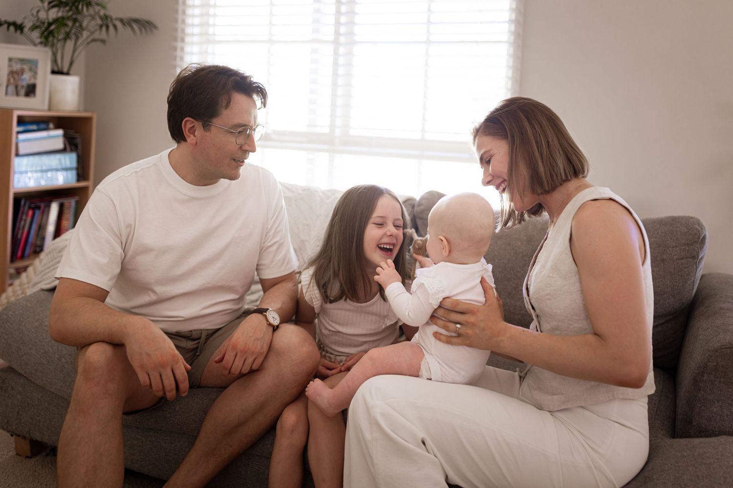 Happy family with baby and young daughter laughing together on couch in cozy living room.
