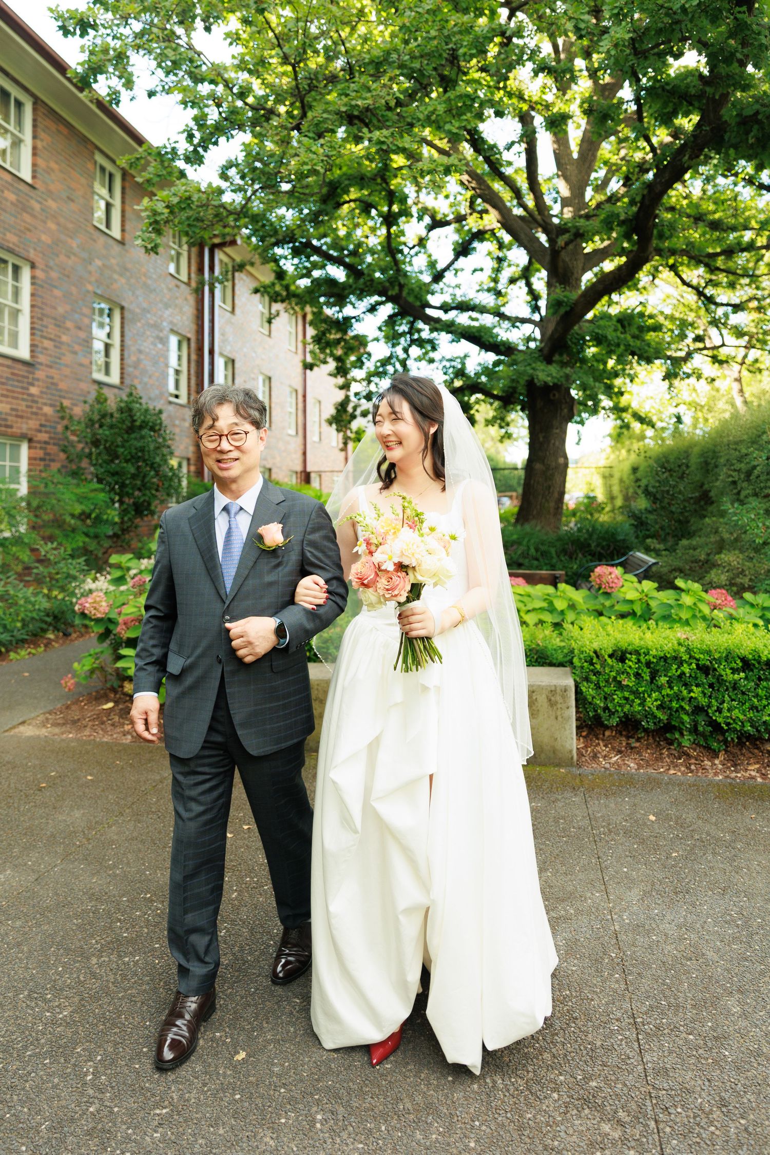 Bride in white gown holding bouquet walking arm in arm with older man in suit outdoors.