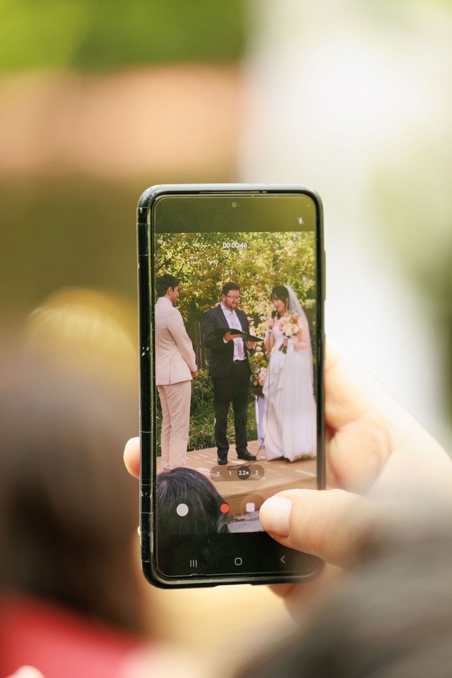 Guest captures wedding ceremony on smartphone, showing couple exchanging vows at outdoor altar.