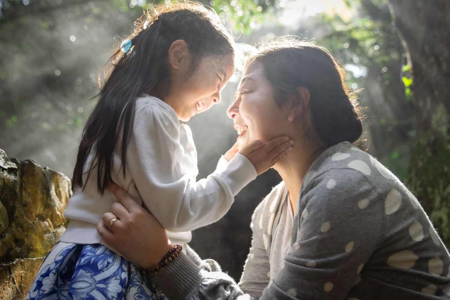 A joyful mother and young daughter share a loving face-to-face moment in soft natural sunlight outdoors.