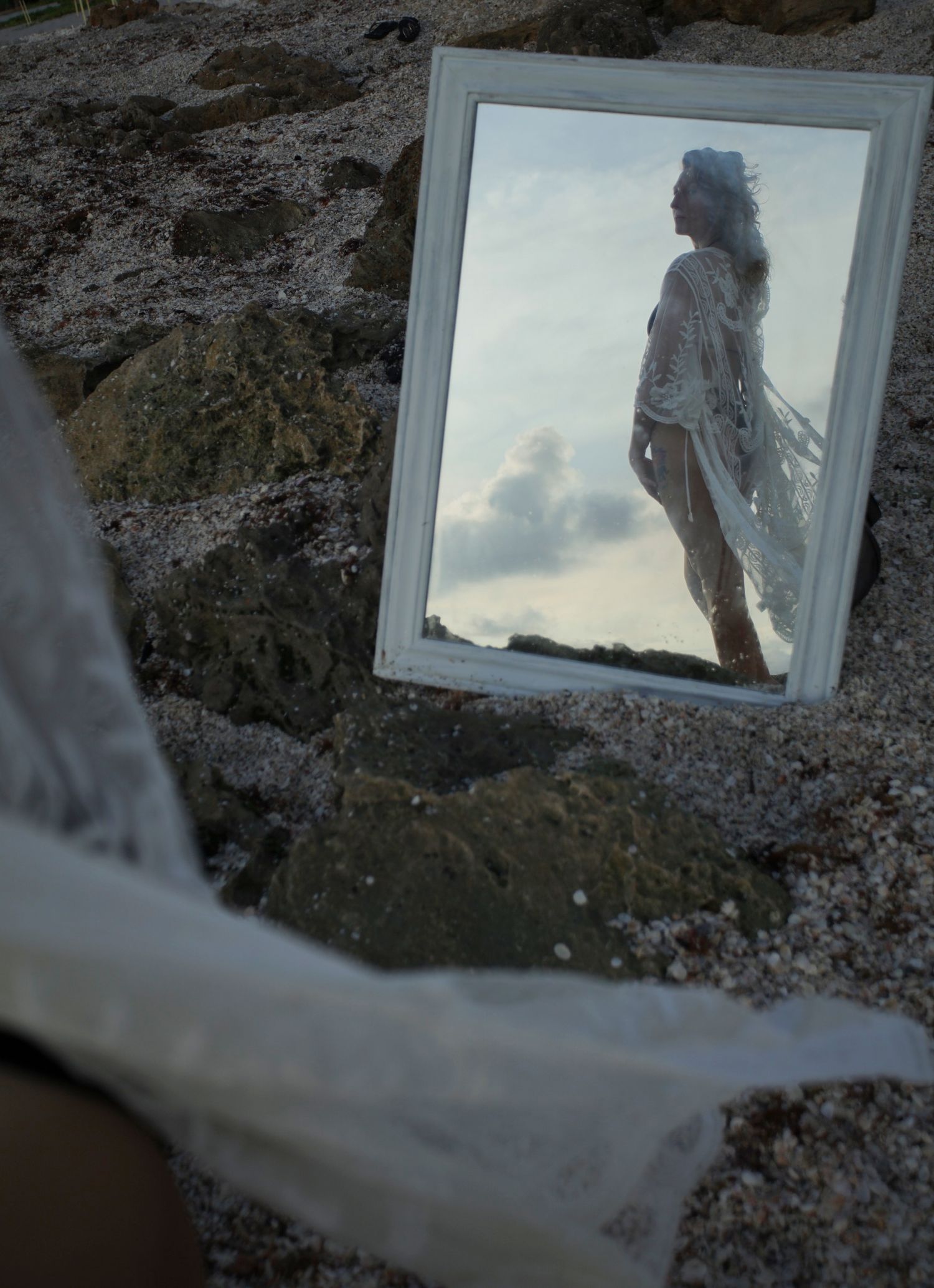 A white framed mirror propped on a rocky beach reflects a woman in a sheer white cover-up against a cloudy sky.