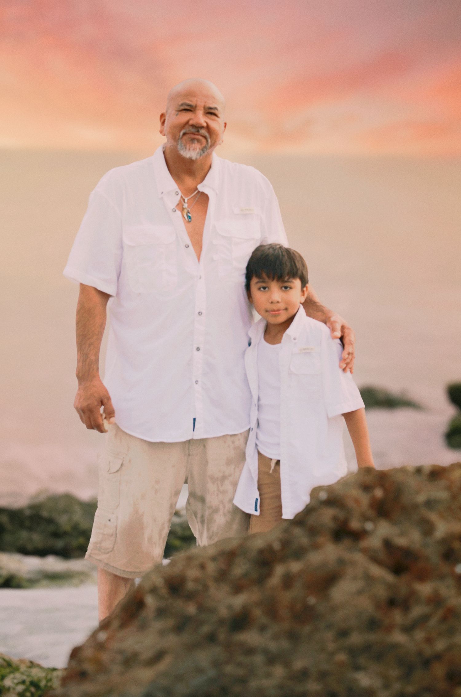 Man and boy in white shirts pose on rocky beach at sunset with pink sky backdrop.