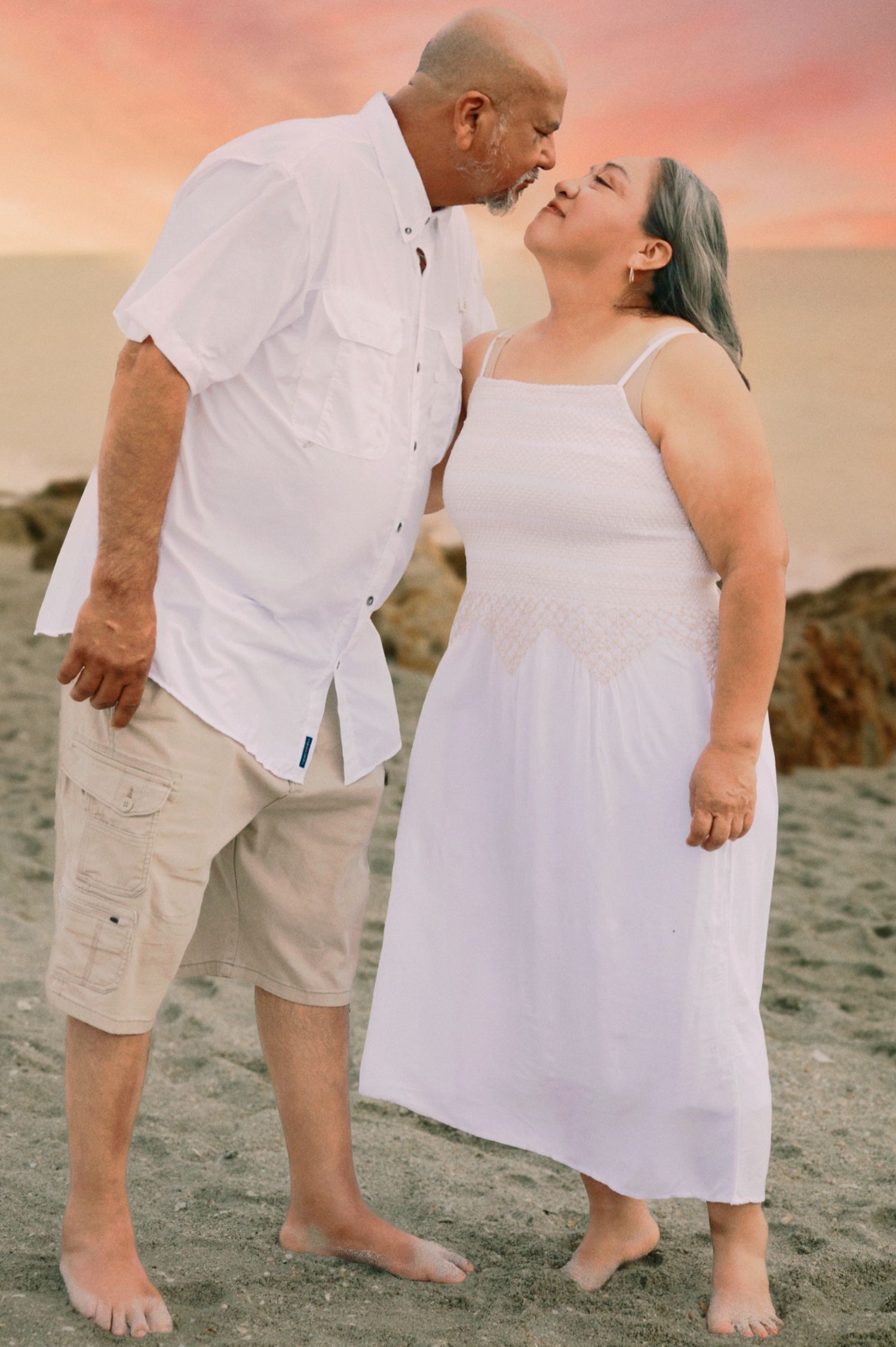 Couple sharing a romantic moment on the beach at sunset, both dressed in white outfits.