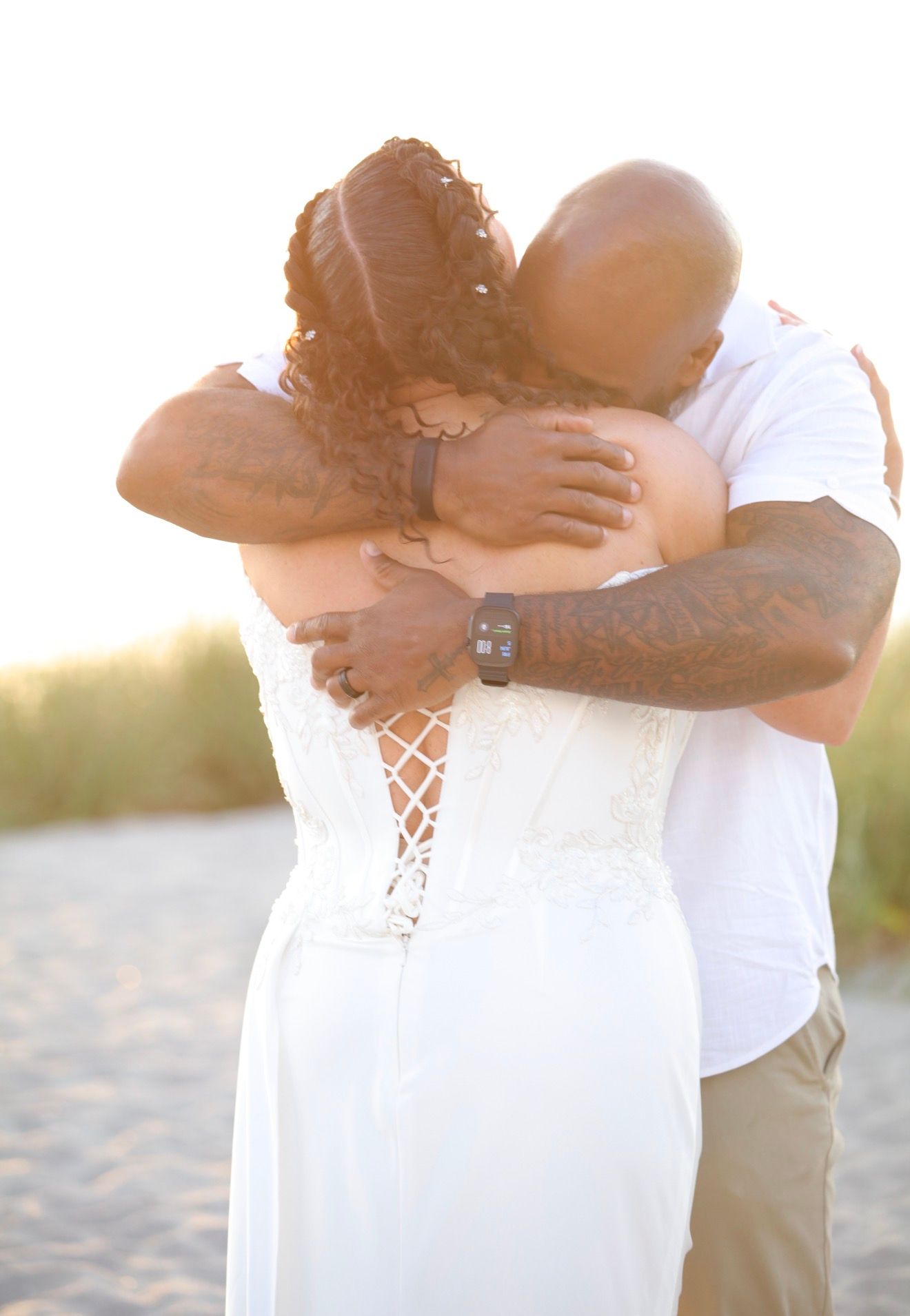 Couple embracing in a tender hug on a sandy beach, woman in white lace wedding dress, golden hour lighting.