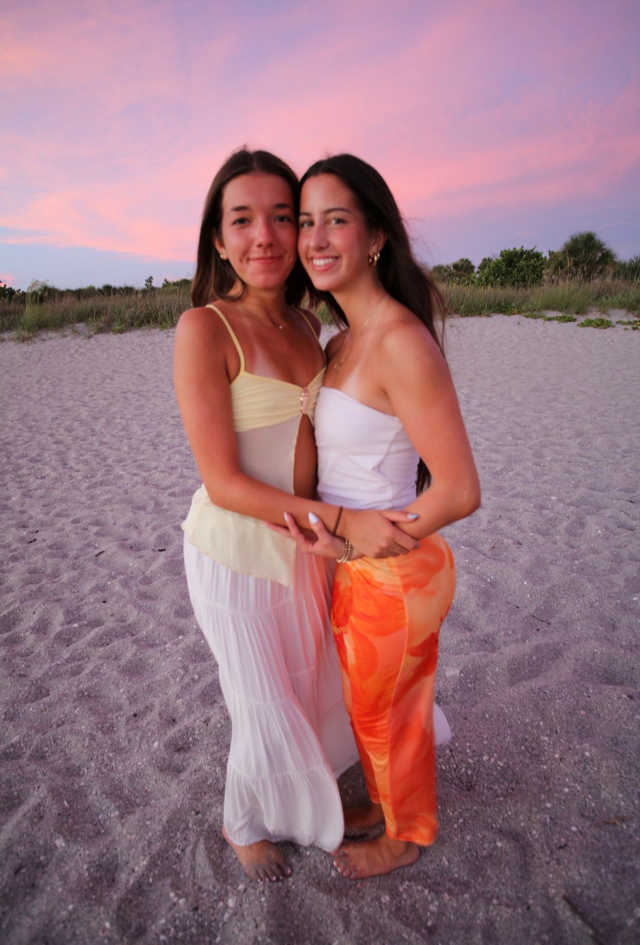 Two young women pose together on a sandy beach at sunset wearing summer dresses with a colorful pink sky.