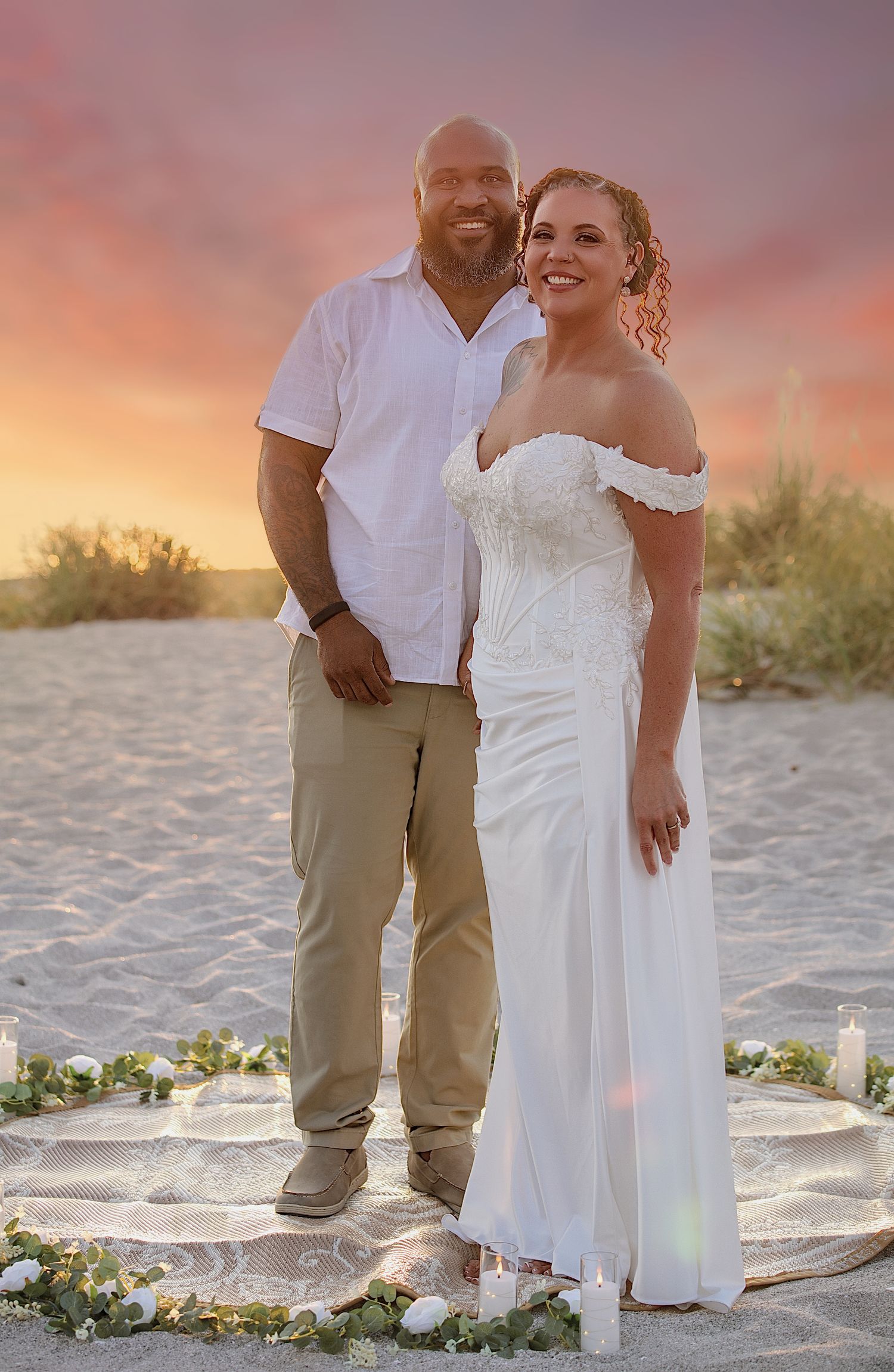 Couple poses on sandy beach at sunset during wedding ceremony, bride in white gown, groom in white shirt and khakis.