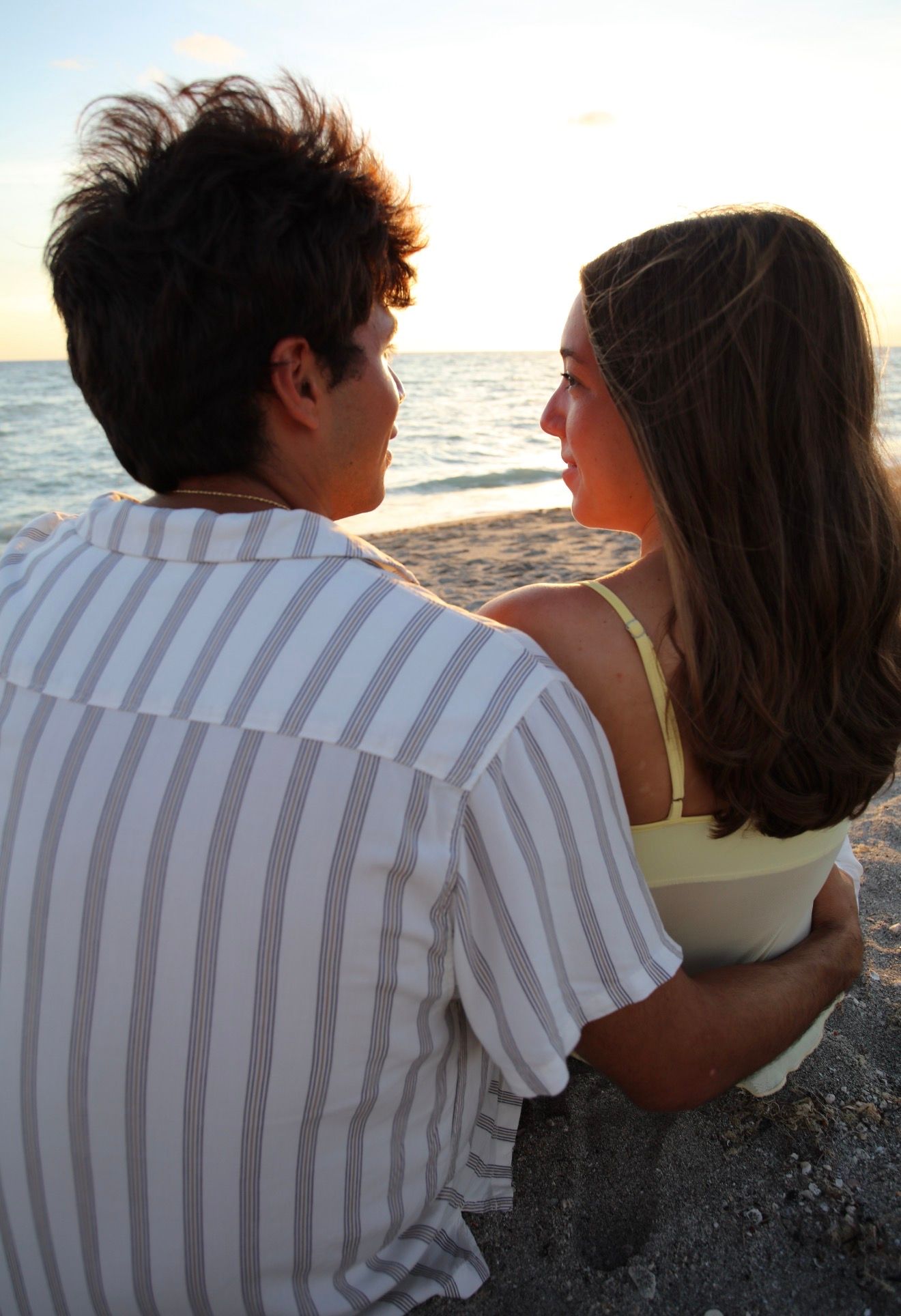 A young couple sits close together on a beach, gazing at each other near the ocean at sunset.