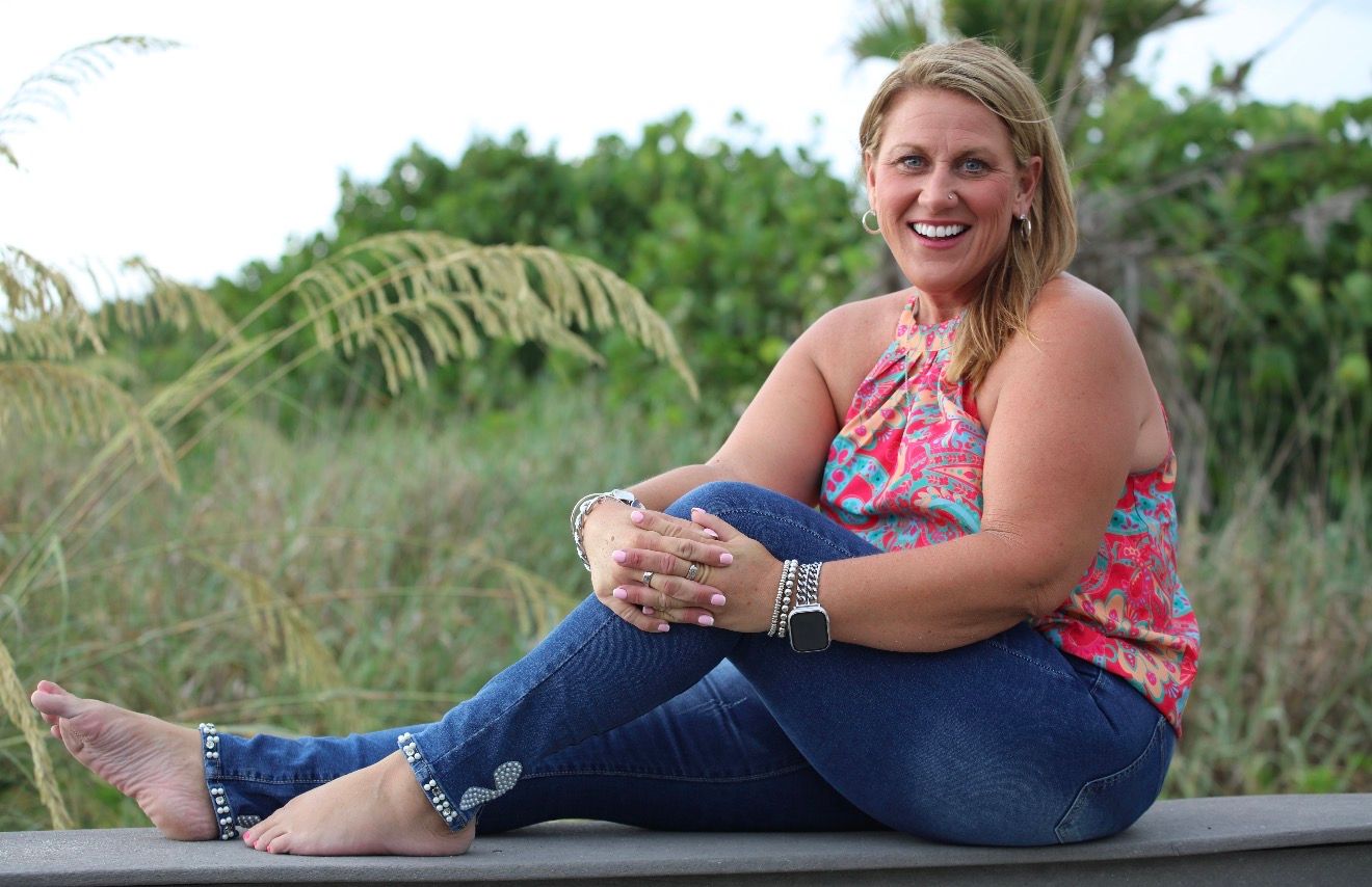 Smiling blonde woman in pink floral top and jeans sitting on wooden deck near coastal greenery.