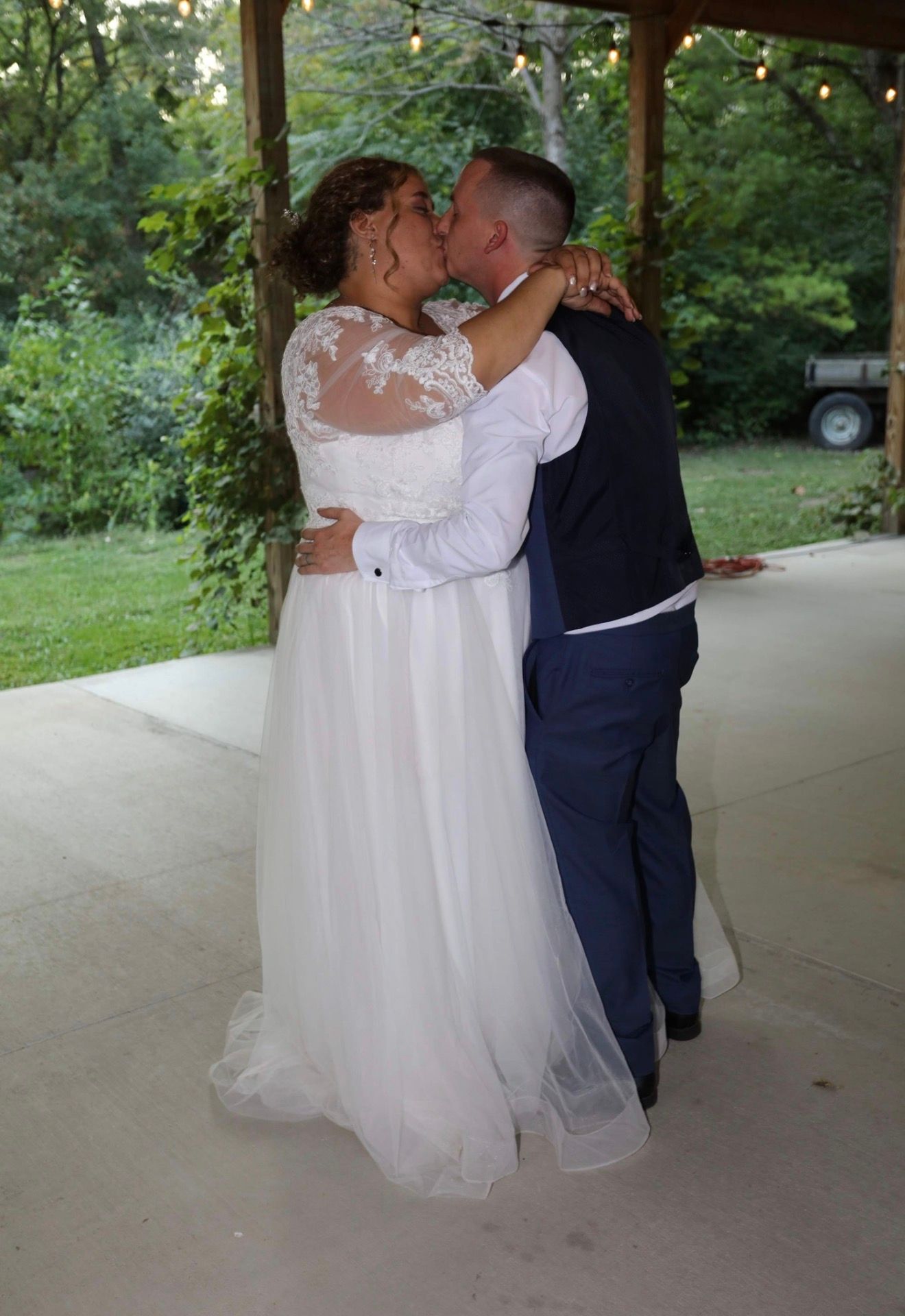Bride and groom share a first kiss in white gown and navy suit under a rustic outdoor pavilion surrounded by greenery.