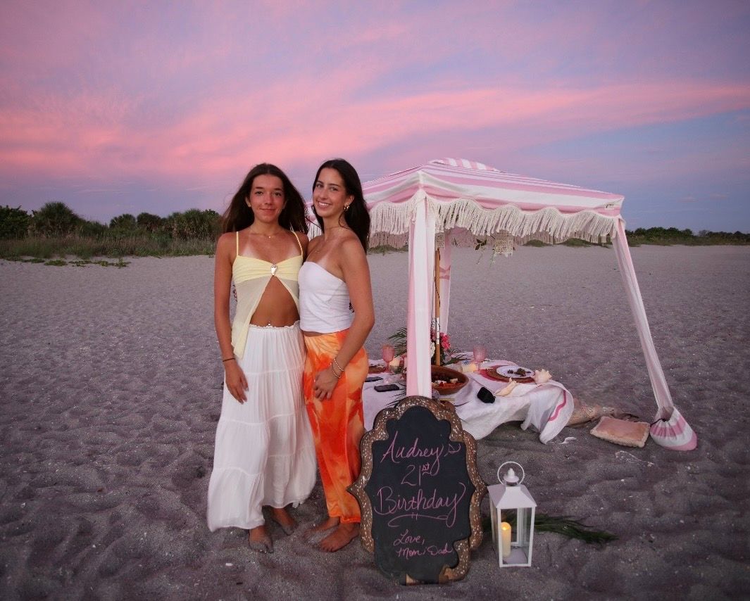 Two women pose on a beach at sunset near a decorated canopy setup for a birthday celebration.