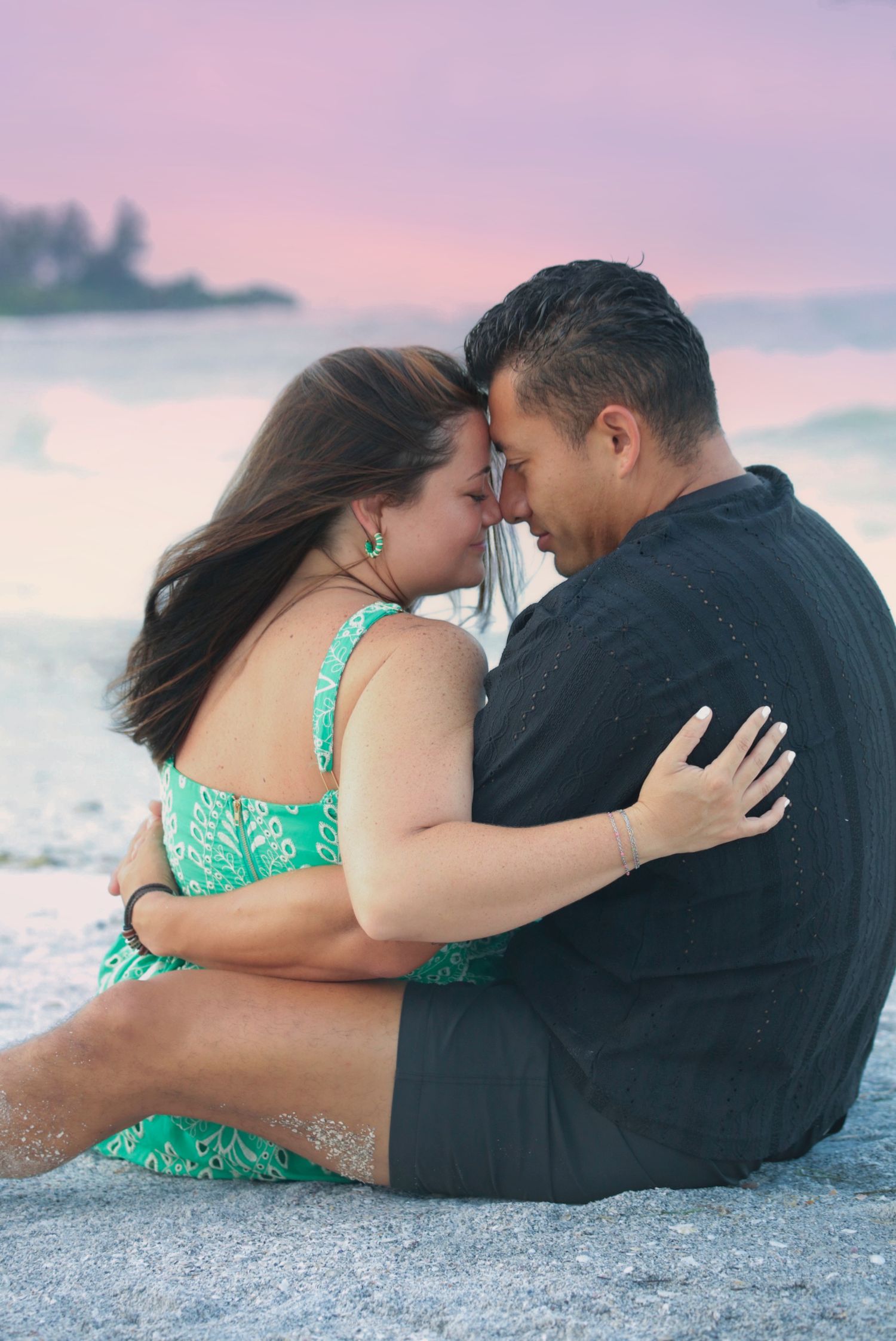 Romantic couple sitting on a sandy beach at sunset, touching foreheads and embracing in an intimate moment.