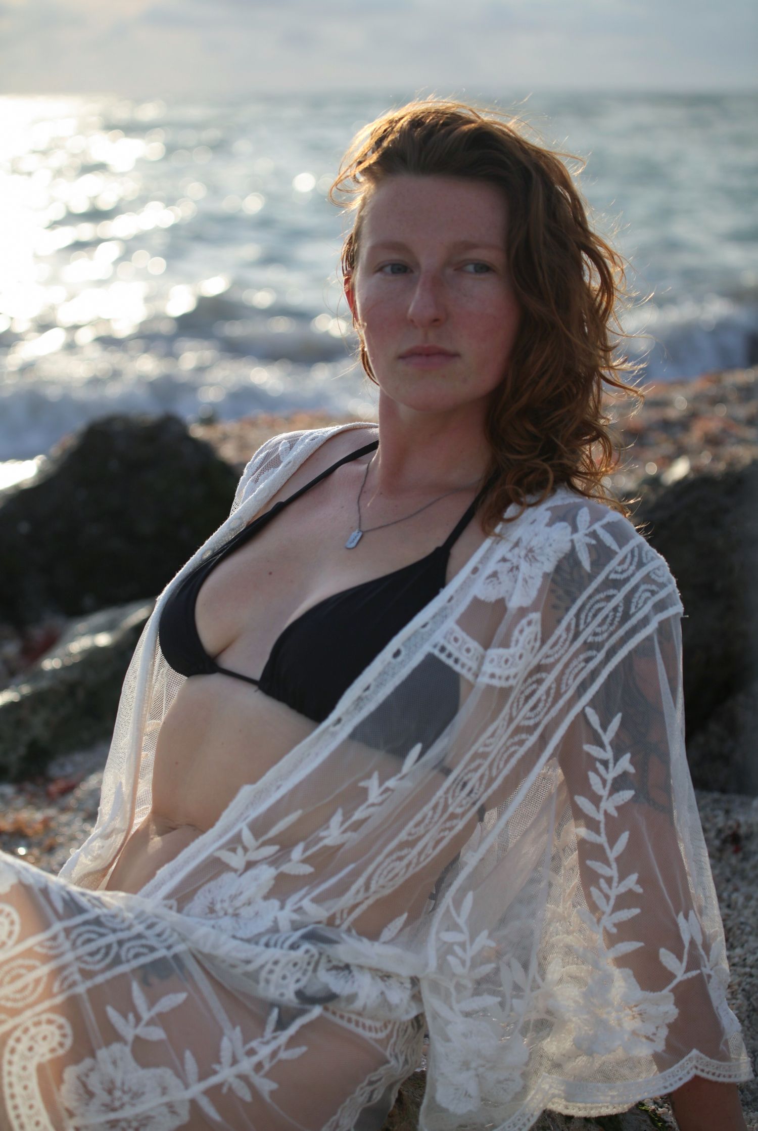 Woman in black bikini and white lace cover-up poses on rocky beach with ocean backdrop.