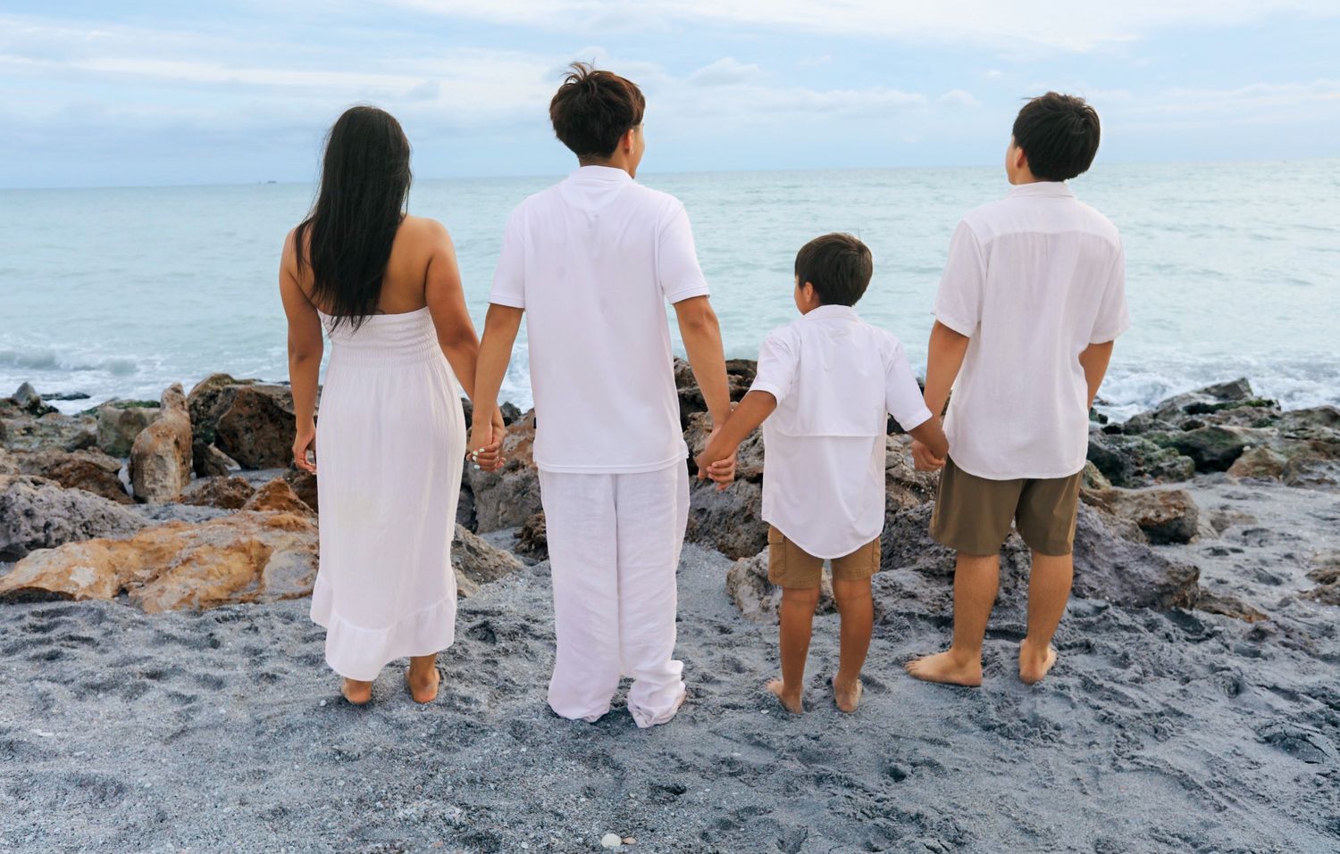 Family of four in white outfits holding hands on rocky beach, viewed from behind with ocean horizon.
