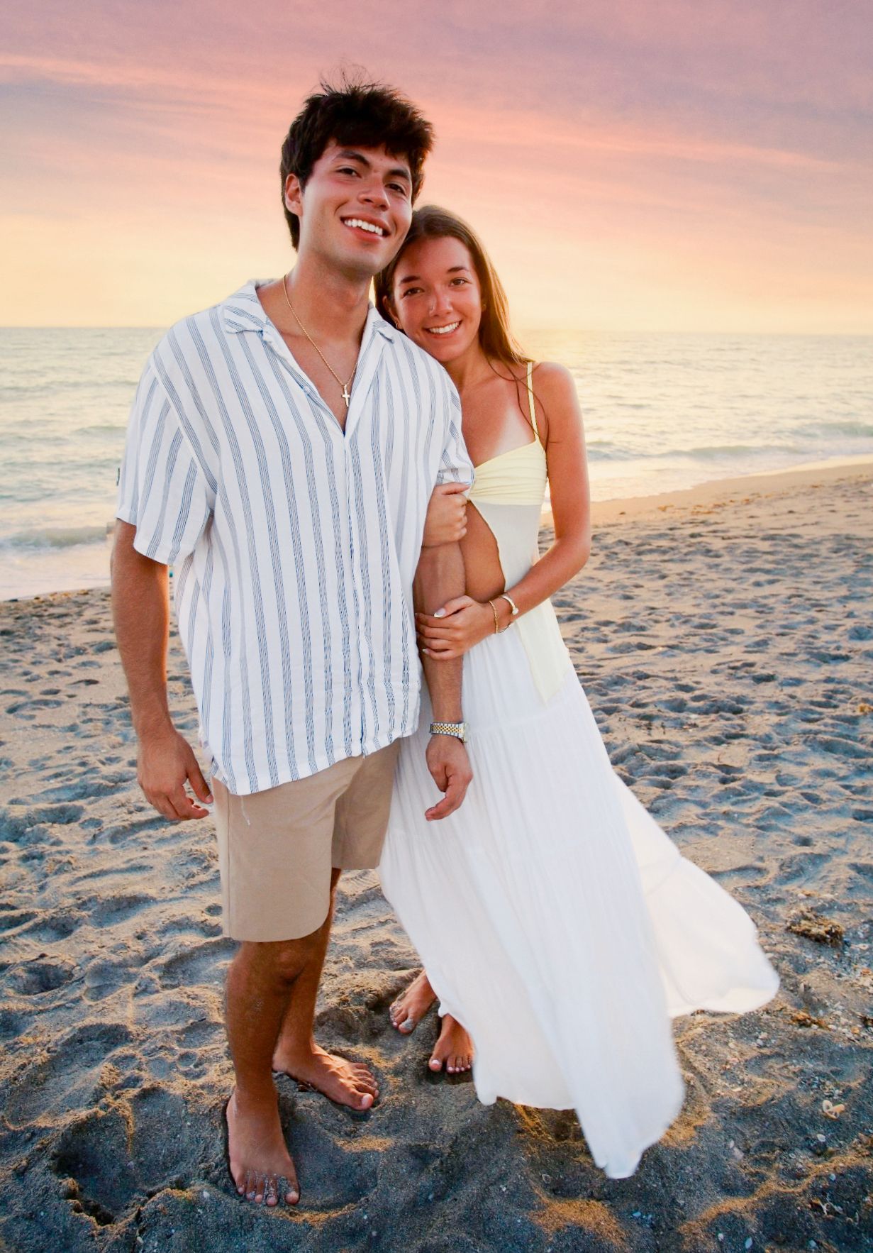 Smiling young couple posing together on sandy beach at pink and purple sunset by the ocean.
